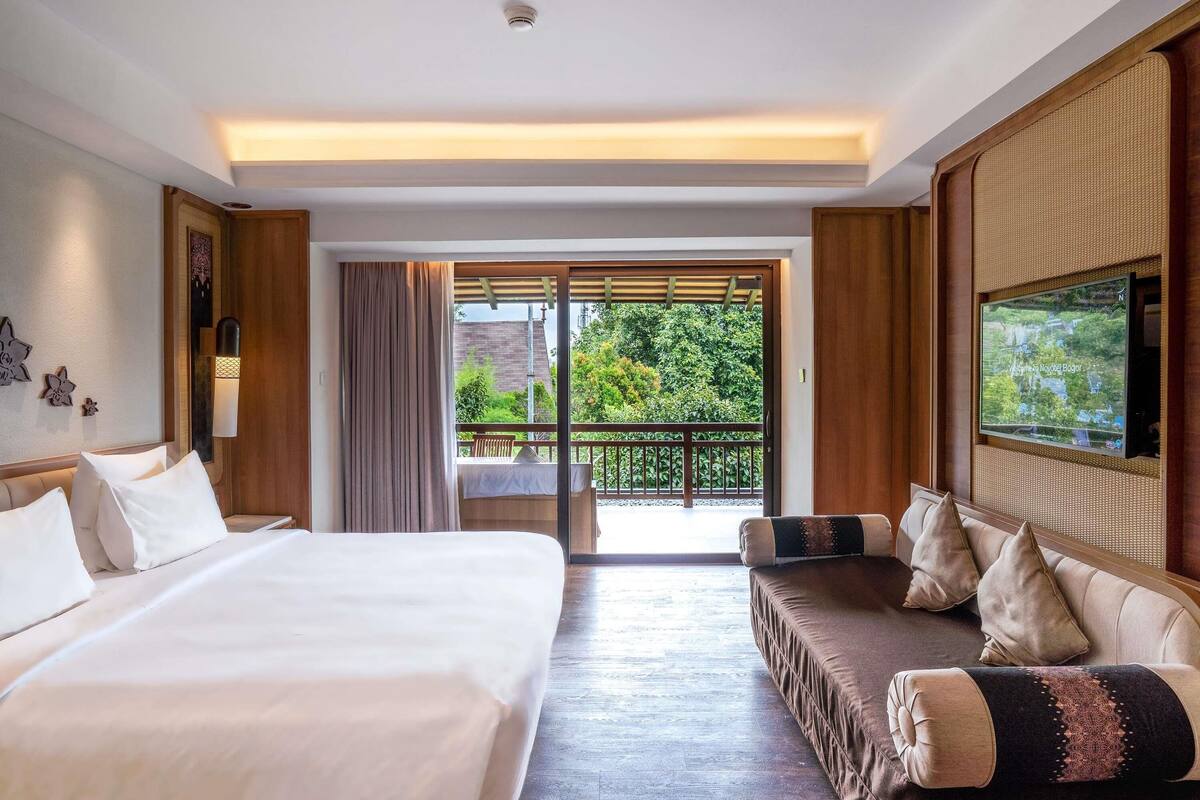 A hotel room with a bed, a sofa, a TV, and a balcony with a view of greenery.