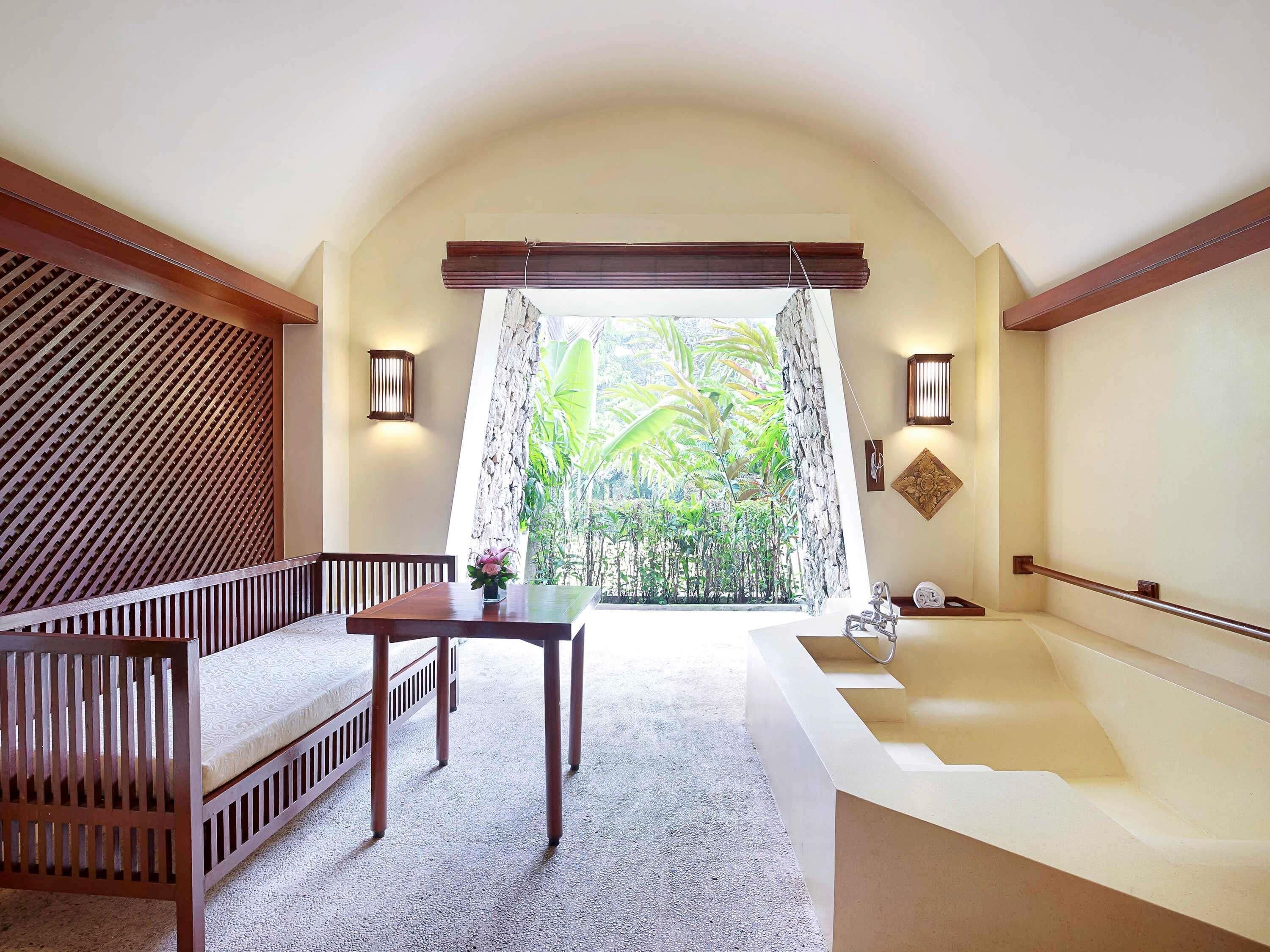 A room with a wooden bench, a table, a bathtub, and a view of greenery through a window.