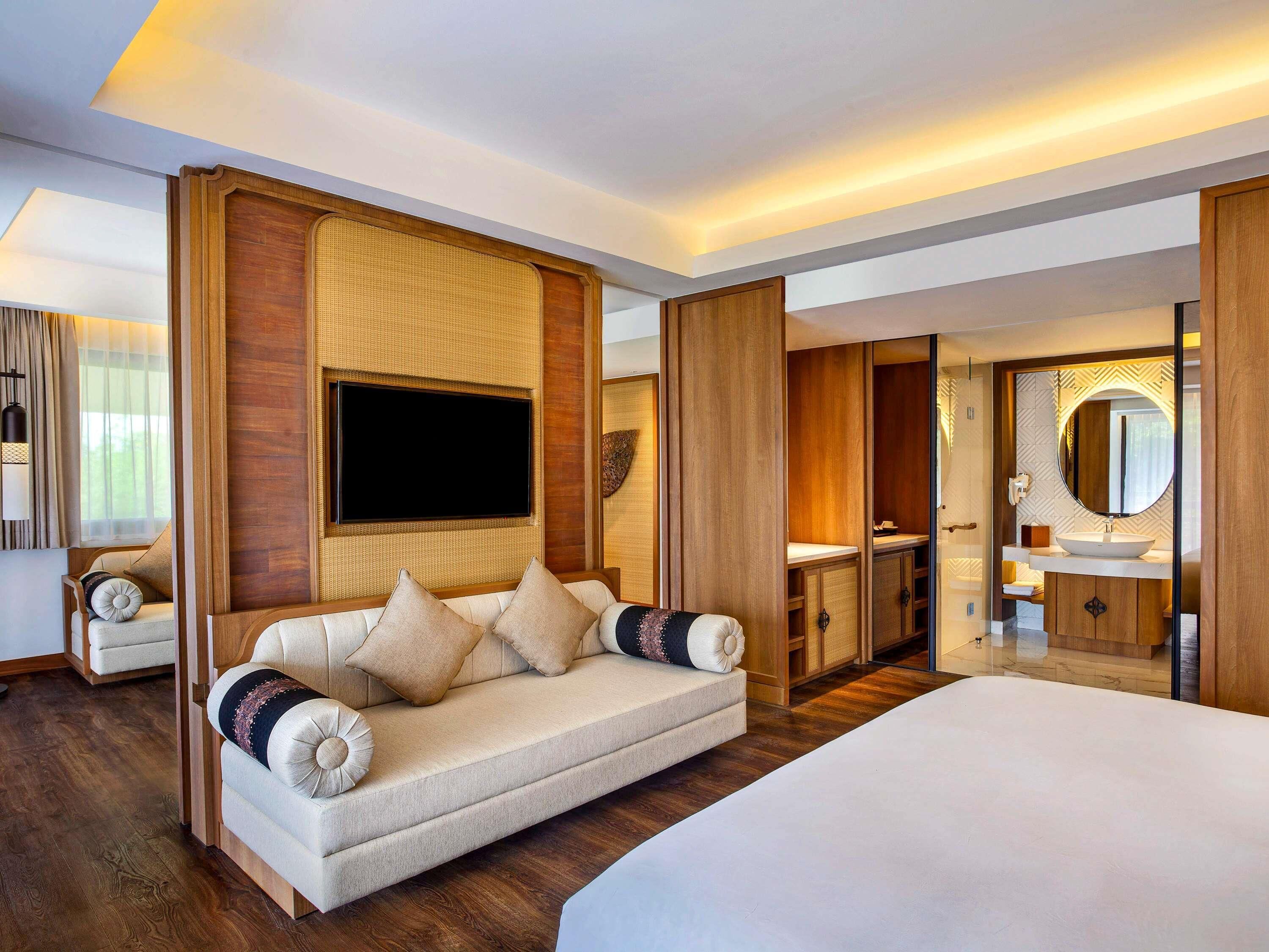 A hotel room with a large bed, a sofa, a TV mounted on a wooden panel, a bathroom with a mirror and sink, and a wardrobe.