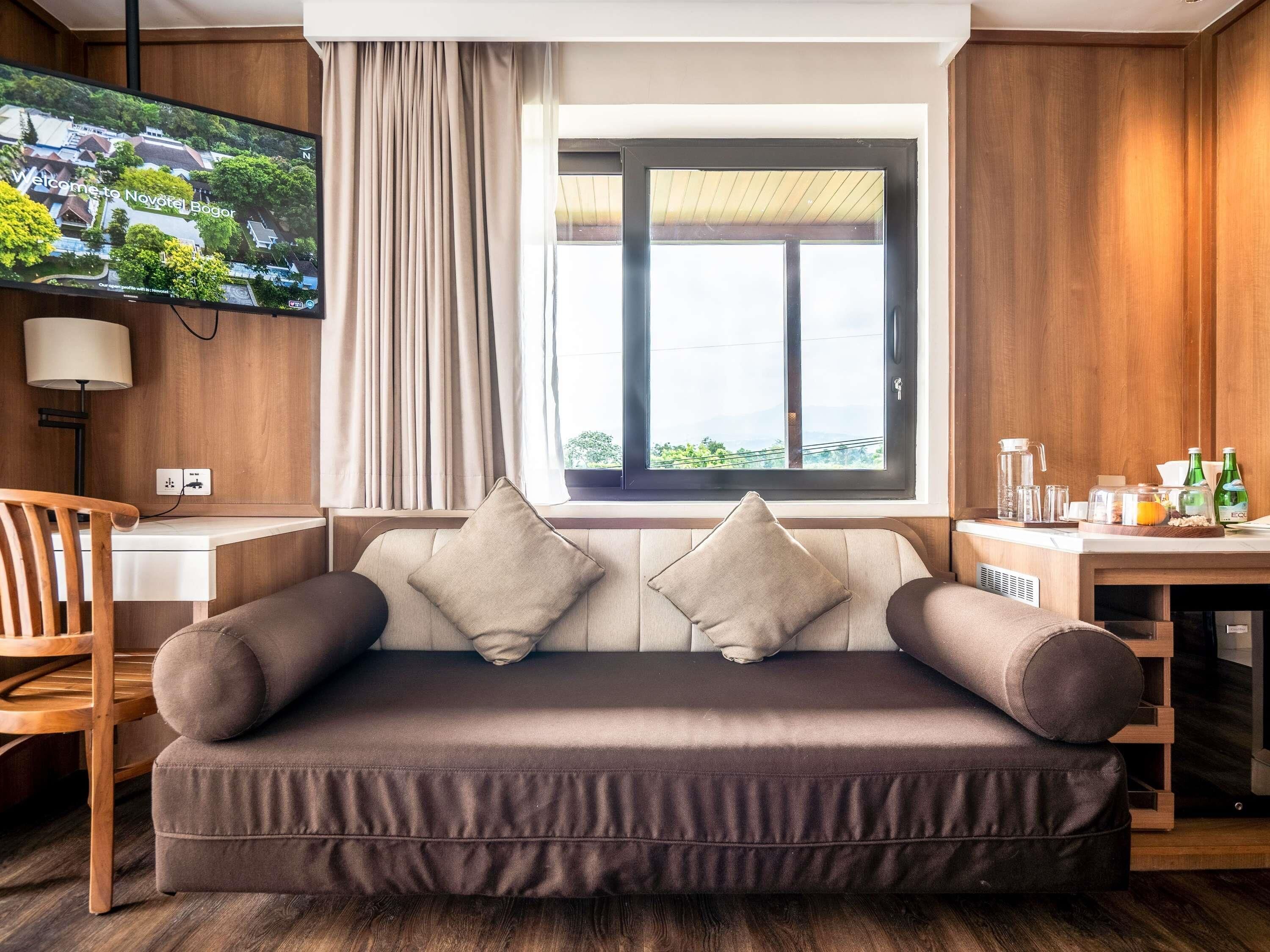A modern living room with a sofa, a TV mounted on the wall, a window with curtains, and a small table with items on it.
