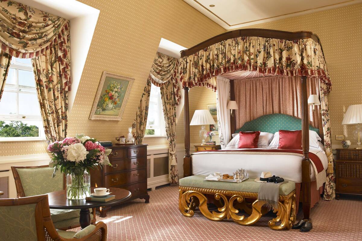 A luxurious bedroom with a four-poster canopy bed, a round table with a vase of flowers, and a view of the outdoors through a window.