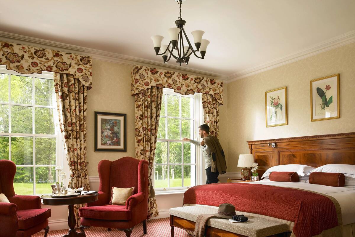 A hotel room with a large bed, a red armchair, a wooden headboard, and a view of the outdoors through large windows.
