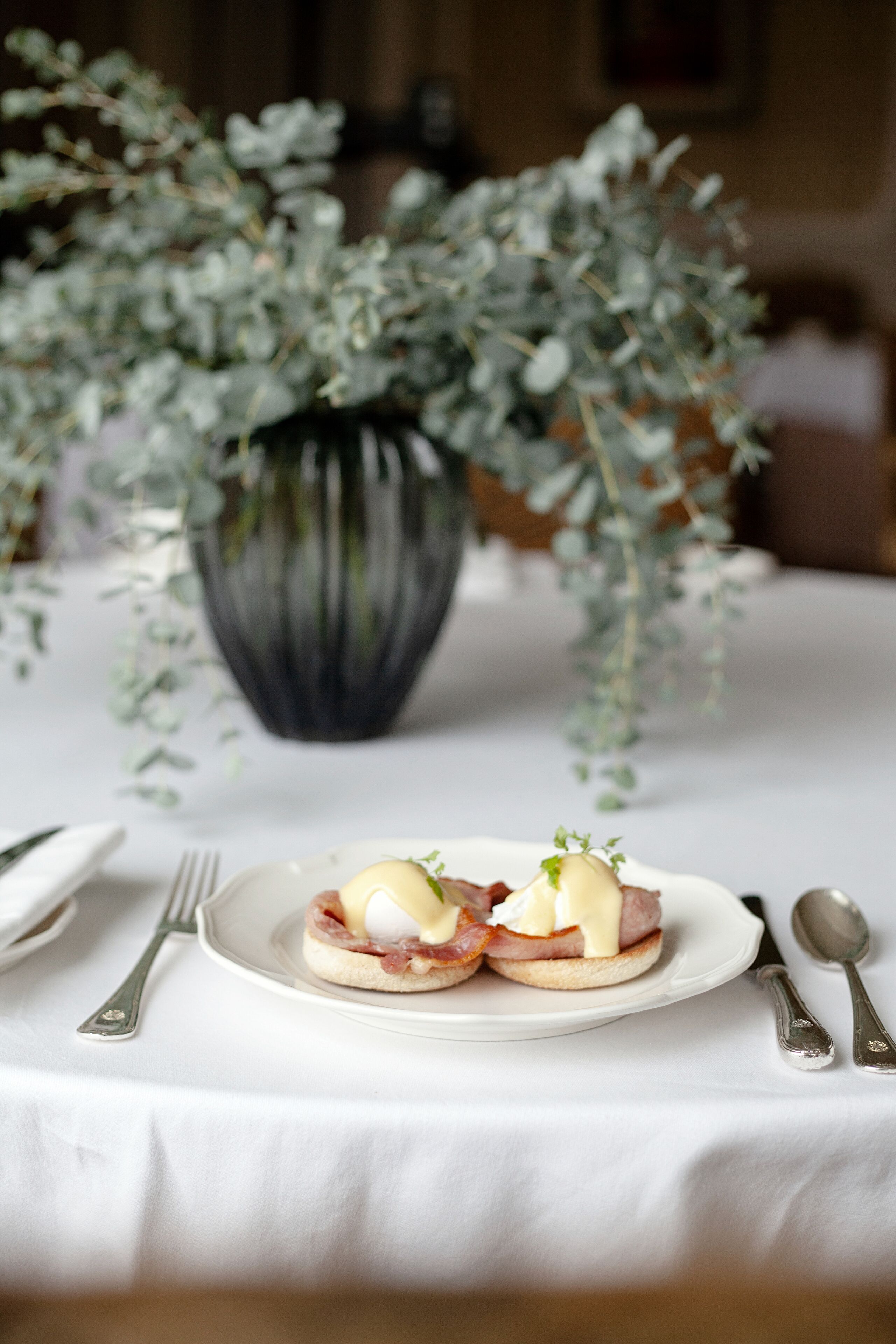 A table with a white tablecloth, a plate with two sandwiches, and a vase with green foliage in the background.