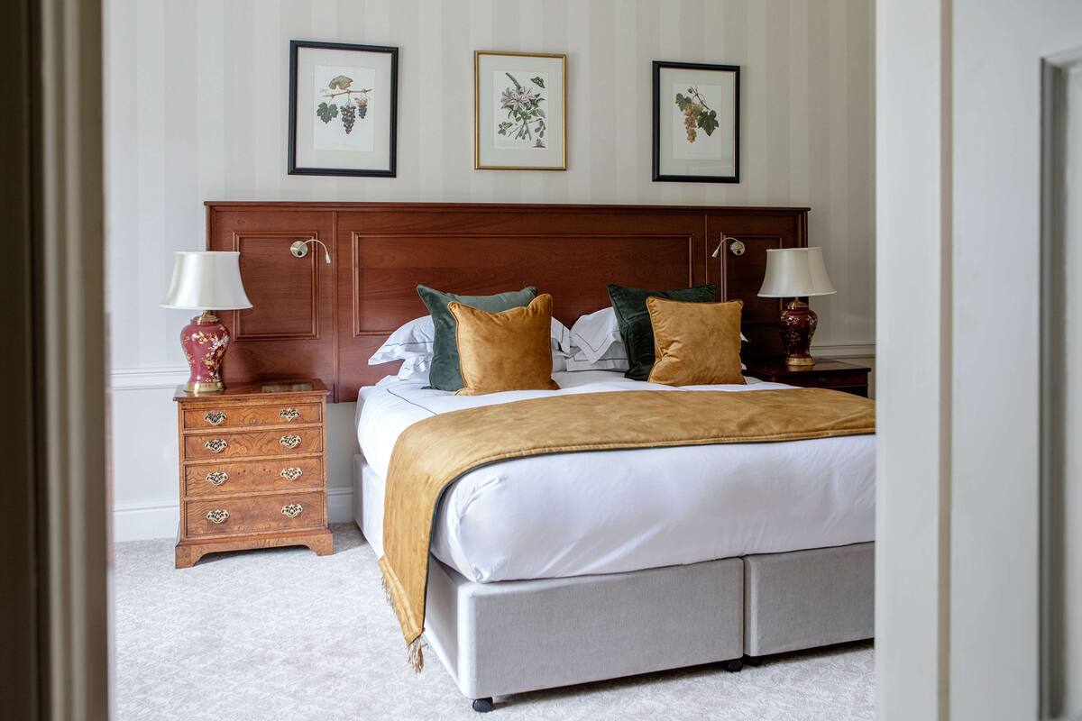 A neatly arranged bedroom with a large bed, two bedside tables with lamps, a wooden dresser, and three framed botanical prints on the wall.