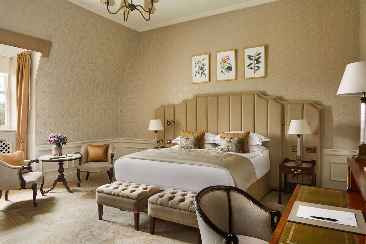 A hotel room with a large bed, two armchairs, a small round table, and a desk with a lamp.