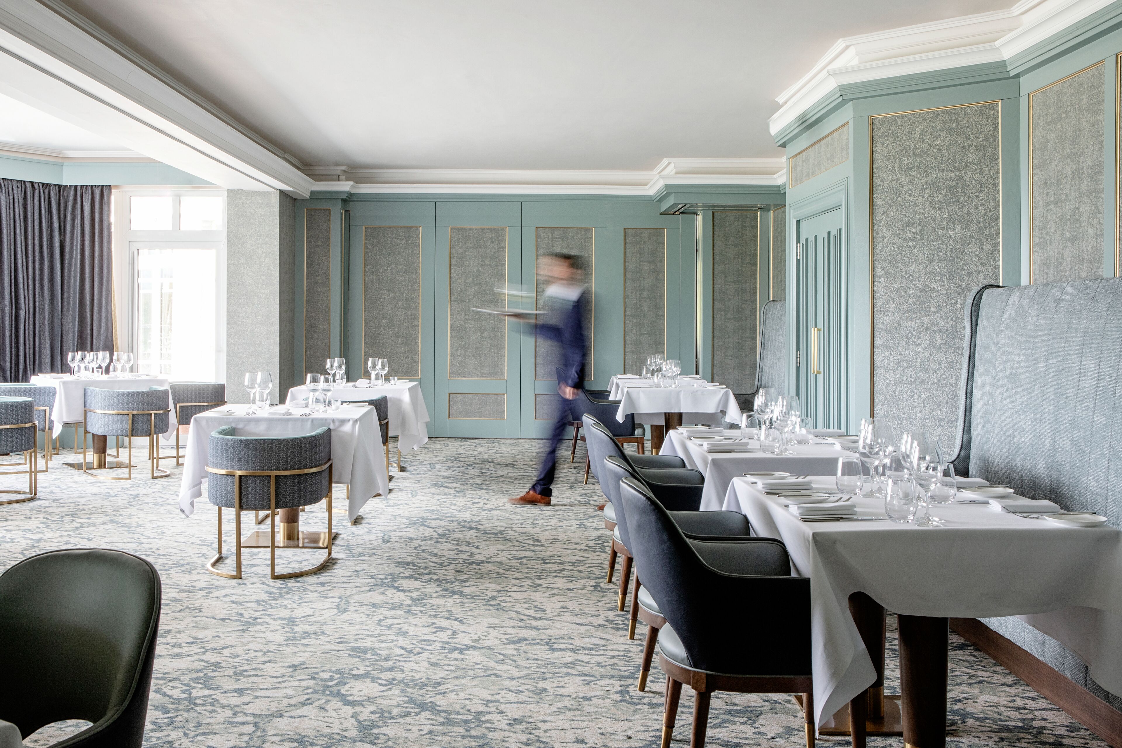 A restaurant with round tables, a waiter in motion, and a patterned carpet.