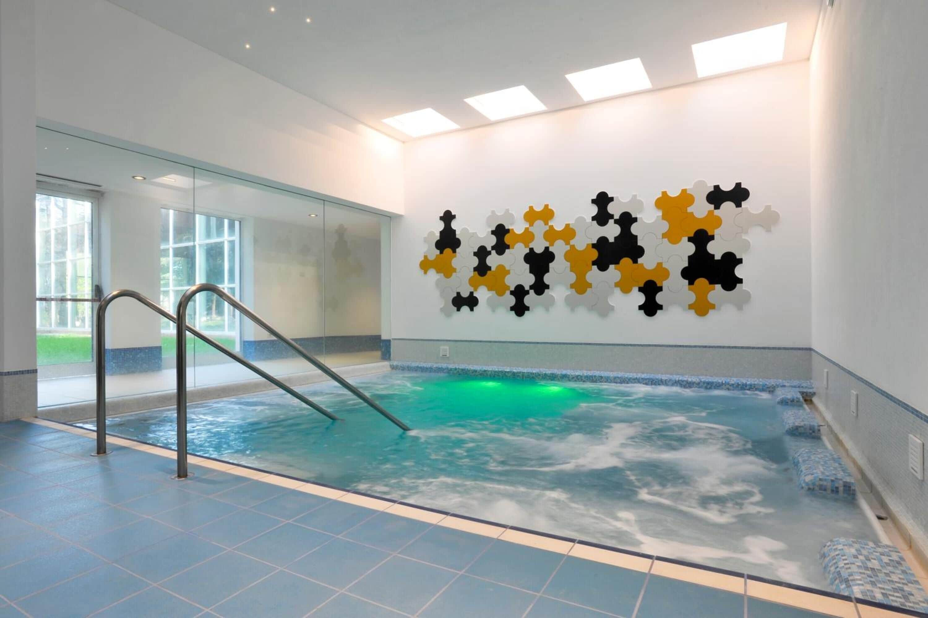 Una piscina interior con pared de vidrio, una escalera metálica e instalación de arte en la pared.