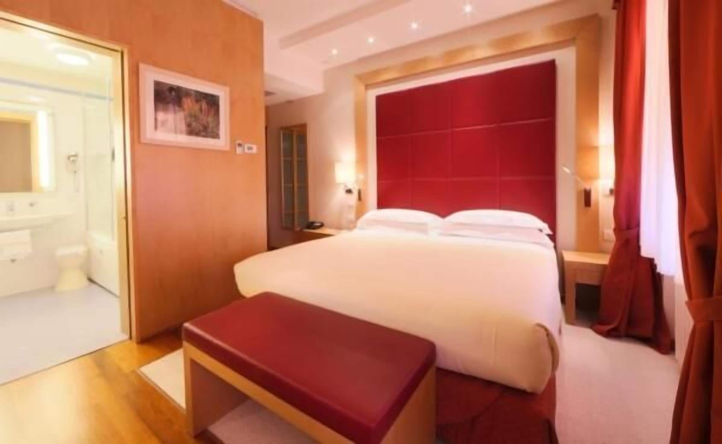 Habitación de hotel con una cama grande, cabecera roja, una mesita de noche pequeña, un banco y un baño visible a través de una puerta abierta.