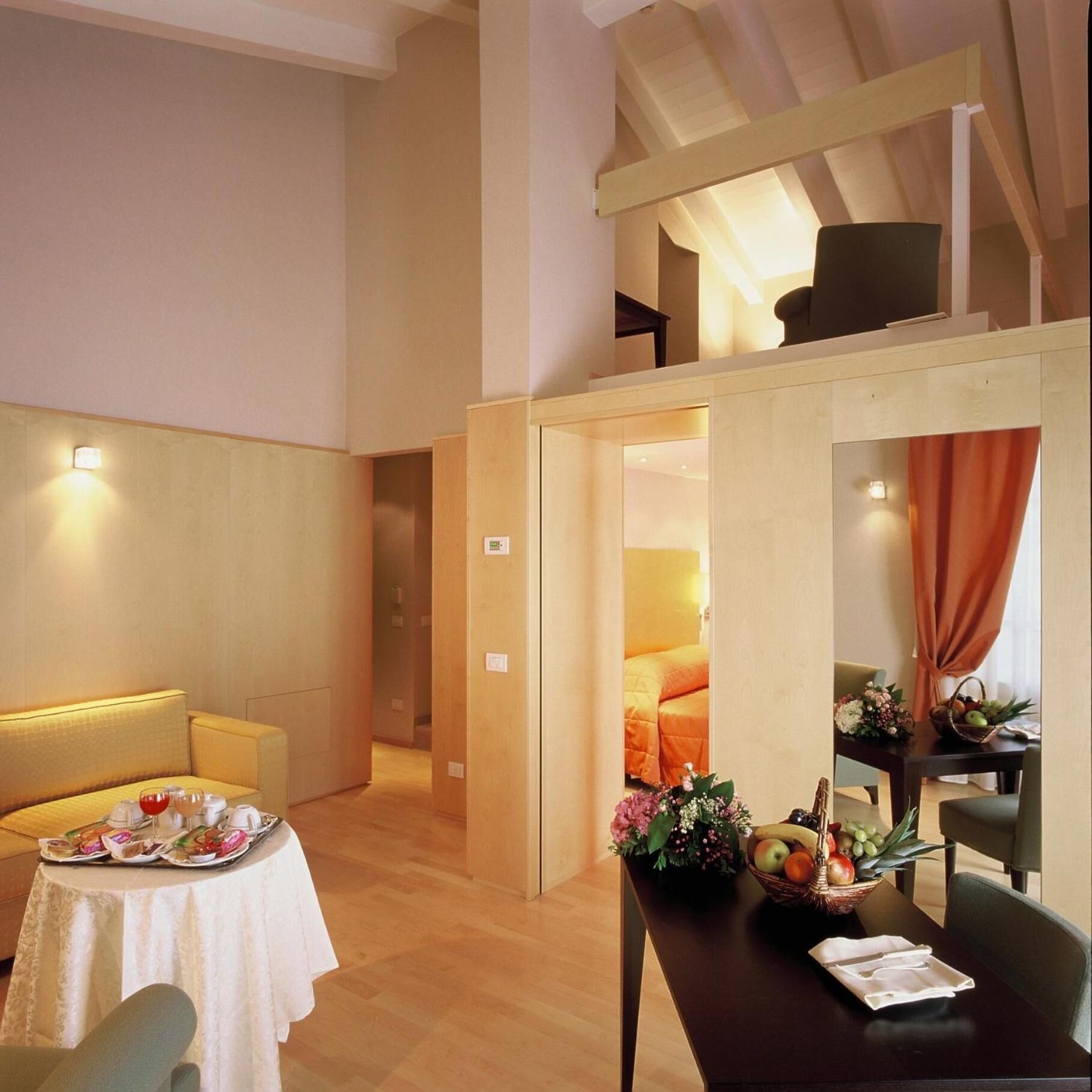 A spacious room with a dining table set for two, a sofa, and a small table with a fruit basket and flowers.