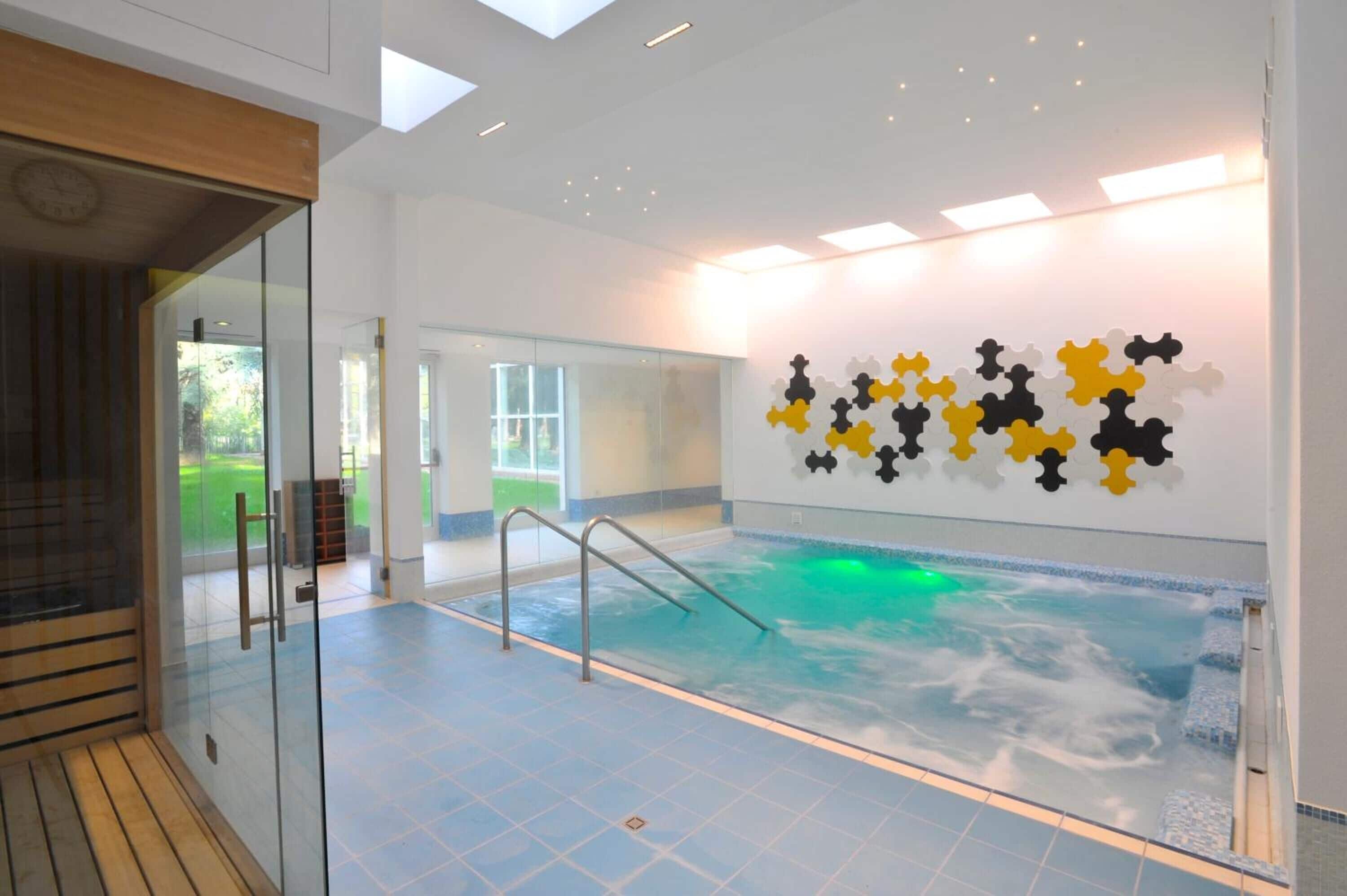 An indoor swimming pool with glass walls, a wooden deck, and a wall art installation.