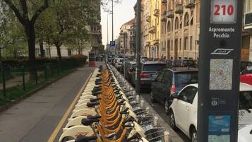 File di stazioni di bike-sharing lungo una strada cittadina, con auto parcheggiate e edifici sullo sfondo.
