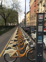 A row of bike-sharing stations along a city street with parked cars and buildings in the background.