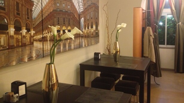 A dining area with a large mural of a historic building, a vase with white flowers, and a table with chairs. - Hotel Aspromonte (Milan)