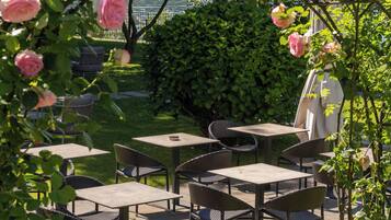 An outdoor patio with tables and chairs, surrounded by lush greenery and blooming pink roses.