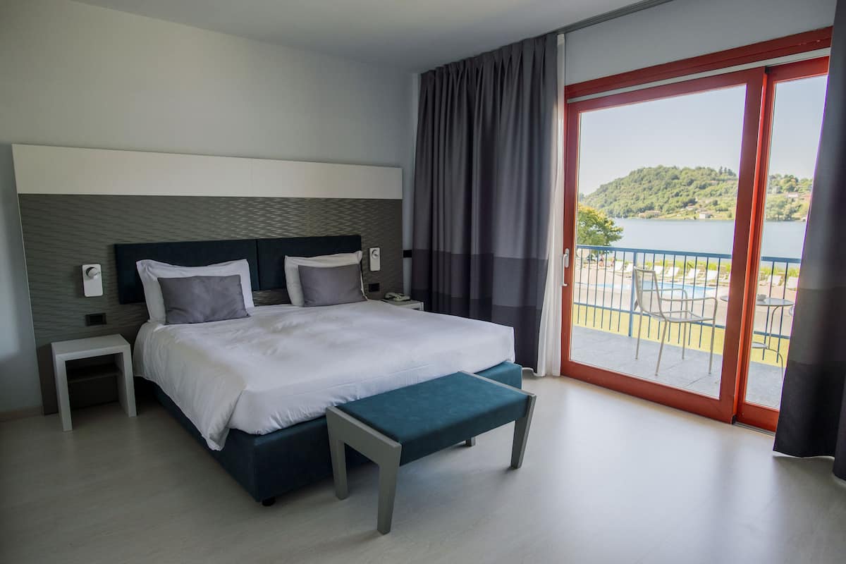 Une chambre d’hôtel moderne avec un grand lit, une banquette, une table de chevet et une vue sur un balcon donnant sur un paysage pittoresque.