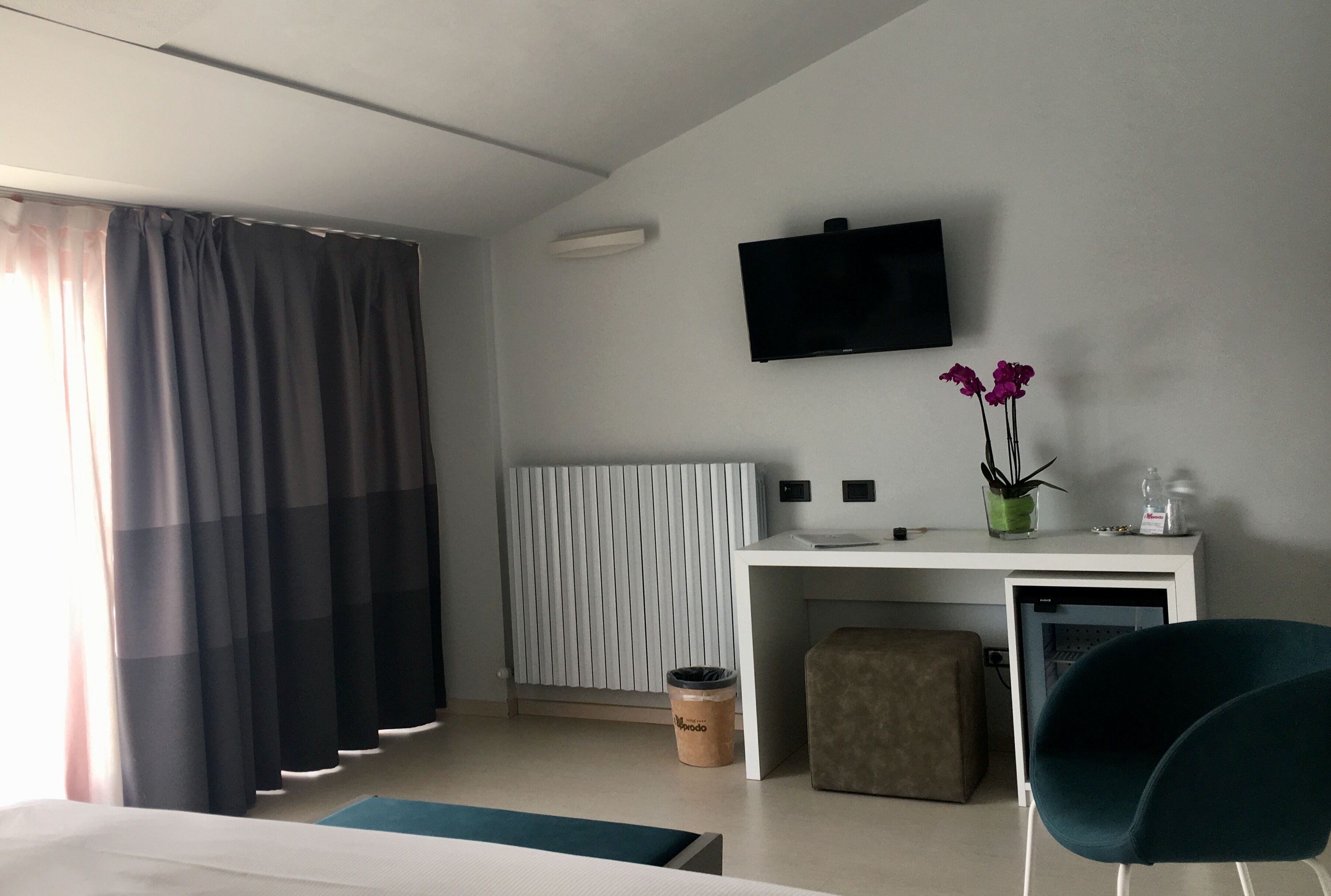 Ein modernes Hotelzimmer mit Bett, Schreibtisch mit Computer, Fernseher, Heizkörper und Mülleimer.