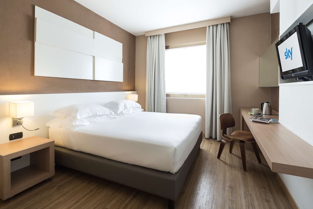 Habitación de hotel con cama, mesita de noche, lámpara, televisor, escritorio, silla y cortinas.