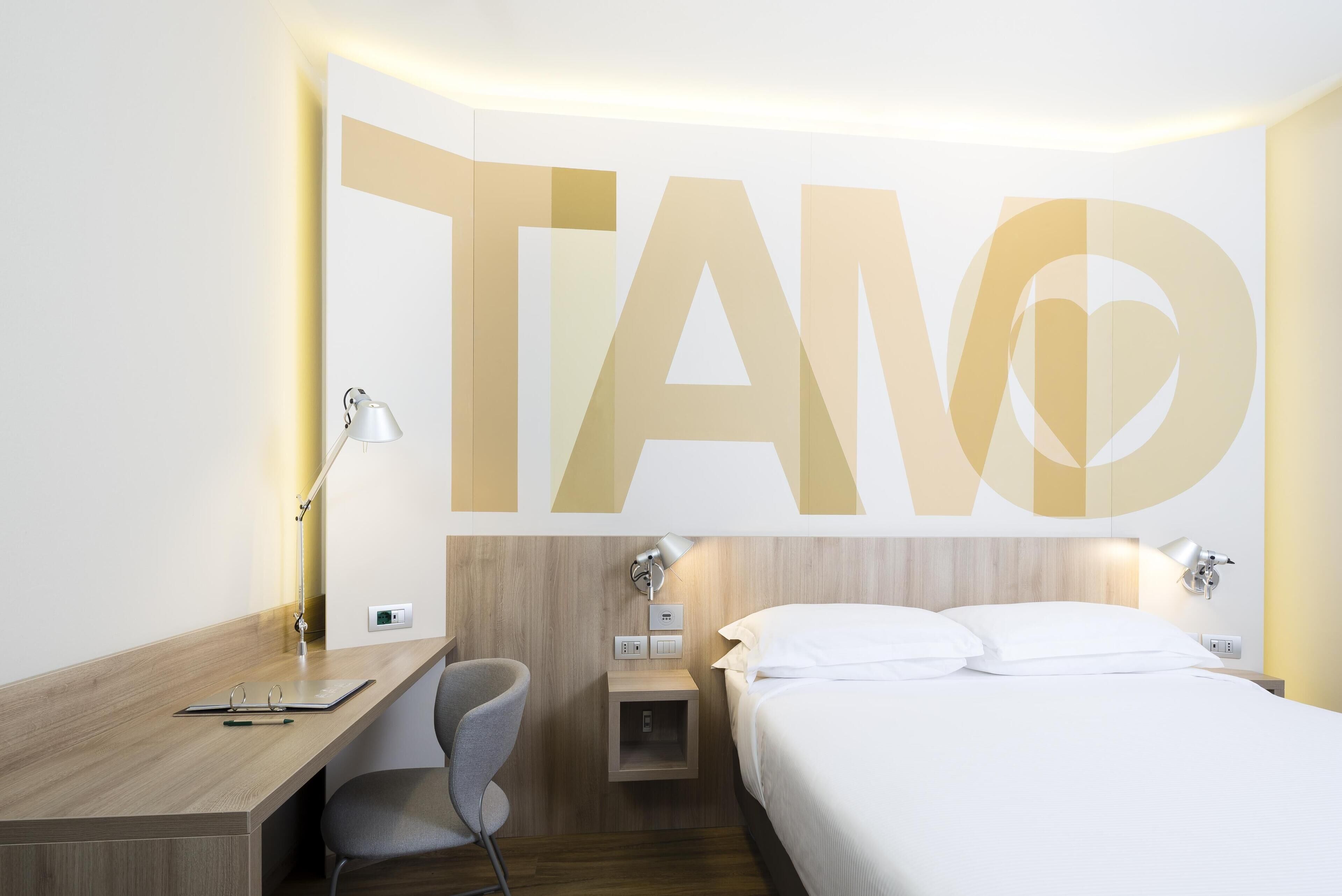 Una camera d'albergo con un letto, una scrivania, una sedia e una parete con la scritta 'TANO' in grandi caratteri.