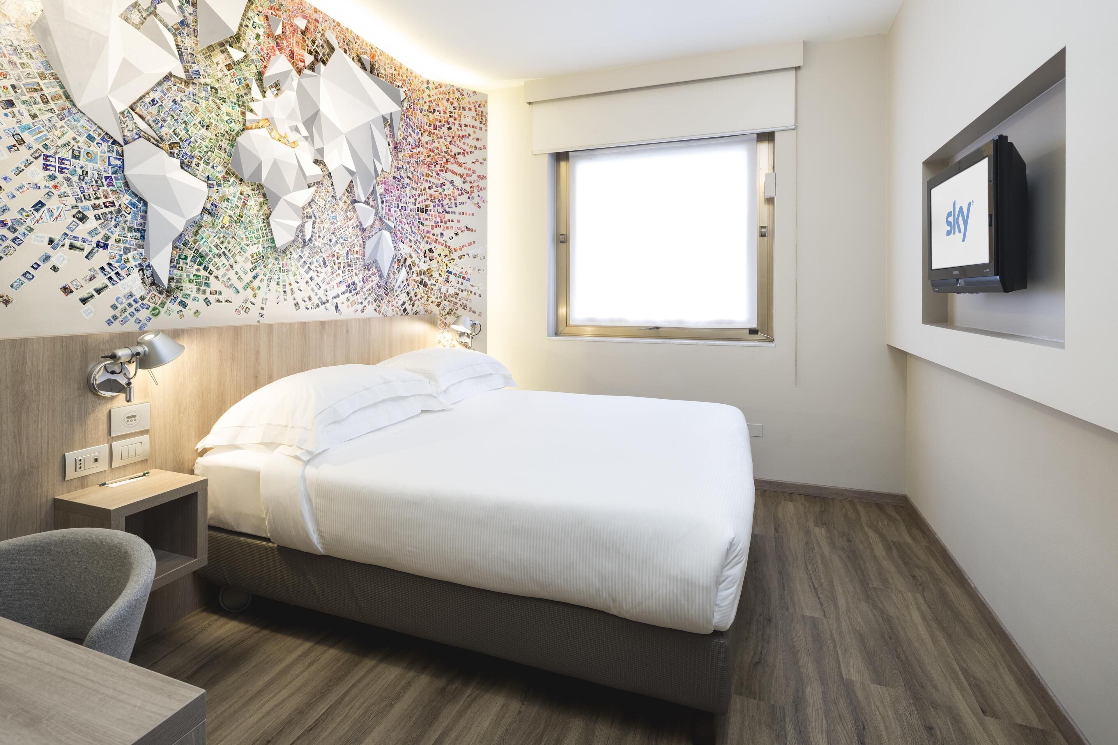 A modern hotel room with a neatly made bed, a desk with a lamp, a wall-mounted TV, and a large window.