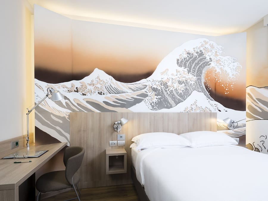 A hotel room with a bed, desk, chair, and a wall mural of a wave.