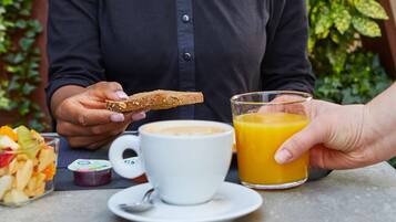 Una persona tiene una fetta di pane tostato e un bicchiere di succo d'arancia a tavola, con una tazza di caffè e una ciotola di macedonia.