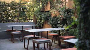 Un patio al aire libre con mesas y bancos de madera, rodeado de follaje y un muro de madera.