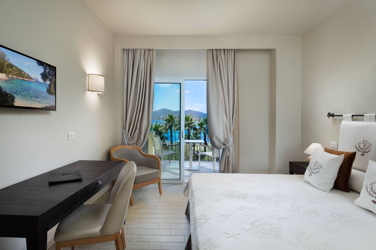 Une chambre d’hôtel équipée d’un lit, d’un bureau, d’une chaise et offrant une vue sur la plage et les montagnes par la fenêtre.