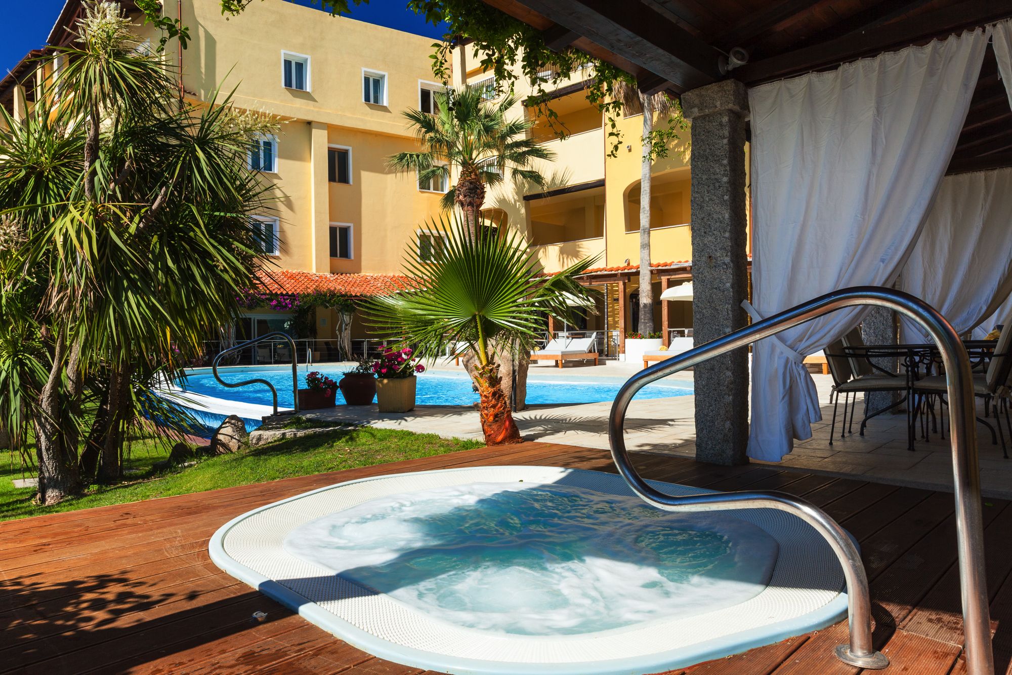 A hot tub with a metal ladder, surrounded by a wooden deck, palm trees, and a pool area with lounge chairs.