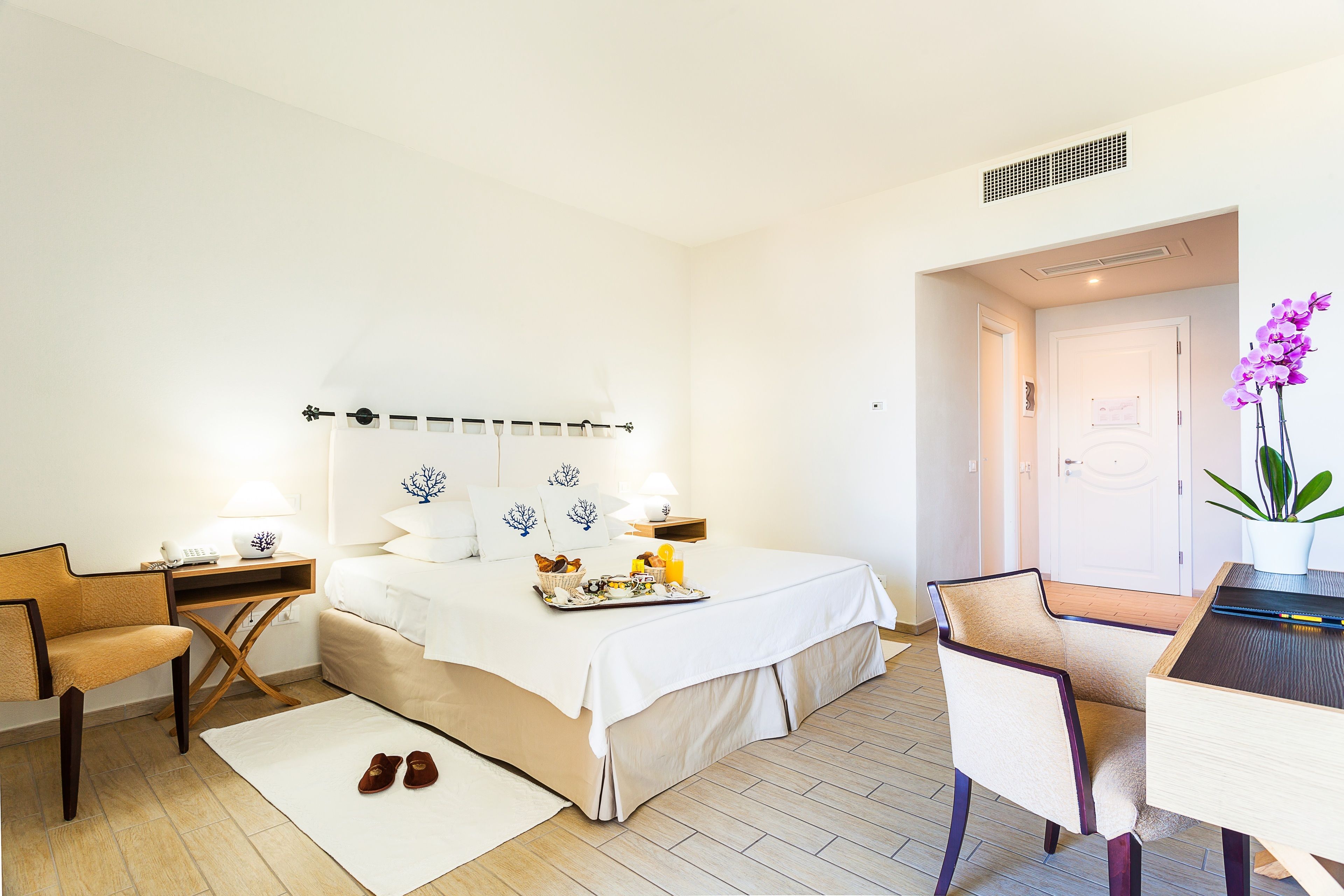 A hotel room with a bed, a desk, a chair, a nightstand, a lamp, a tray of breakfast, and a potted plant.