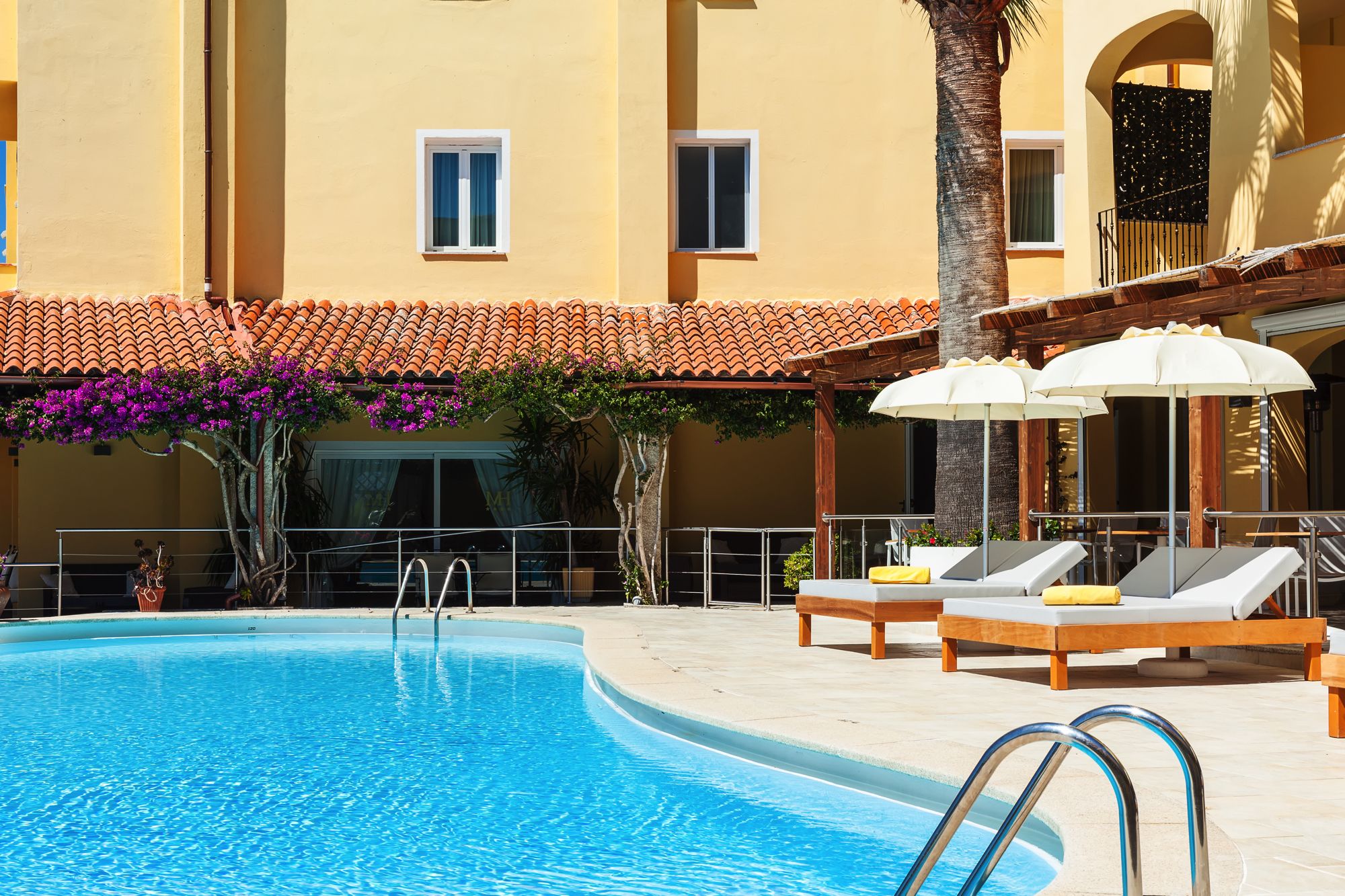Une piscine entourée de chaises longues et de parasols, le tout bordé d’un bâtiment doté d’un toit en tuiles et de balcons.