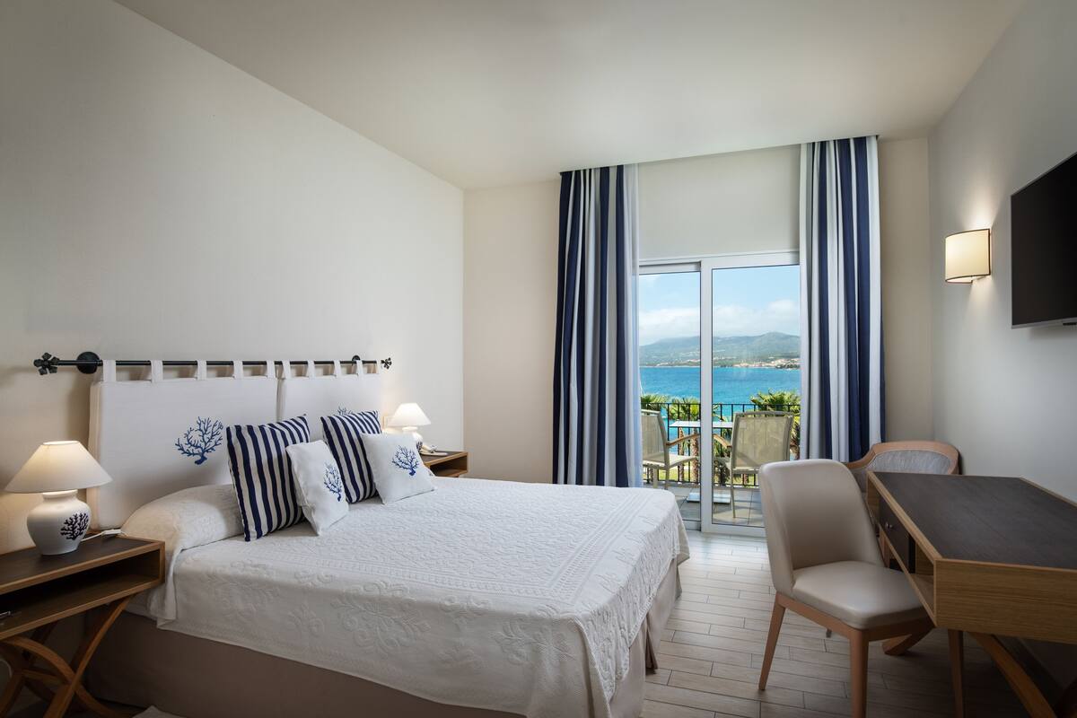 Une chambre d’hôtel avec un lit, une table de chevet, une lampe et un bureau avec une chaise. Il y a une grande fenêtre avec des rideaux et une vue sur la mer.