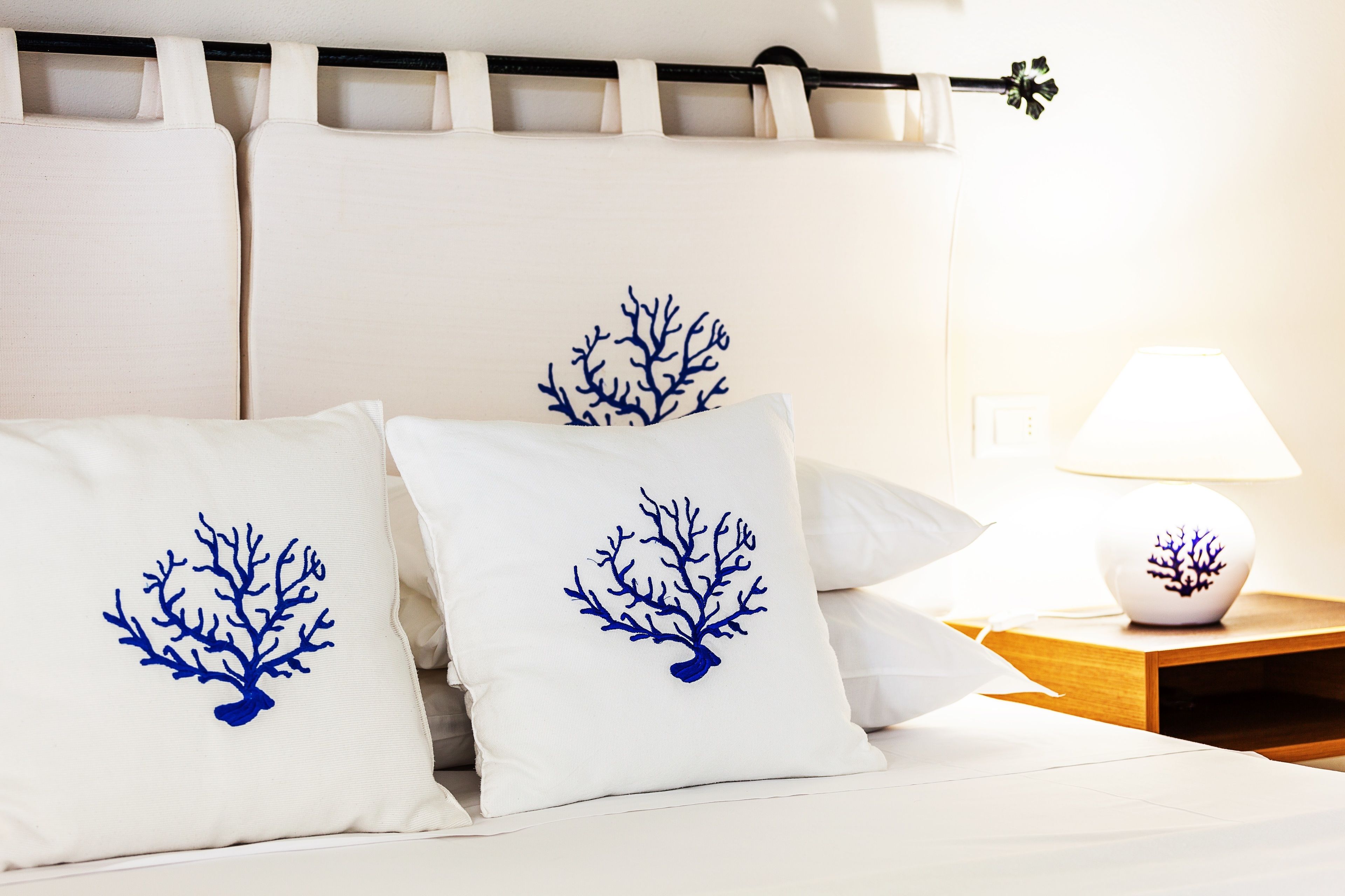 Ein Bett mit weißer Bettwäsche und zwei Kissen mit einem blauen Korallenmuster. Ein Nachttisch mit Lampe und einem kleinen dekorativen Gegenstand ist zu sehen.