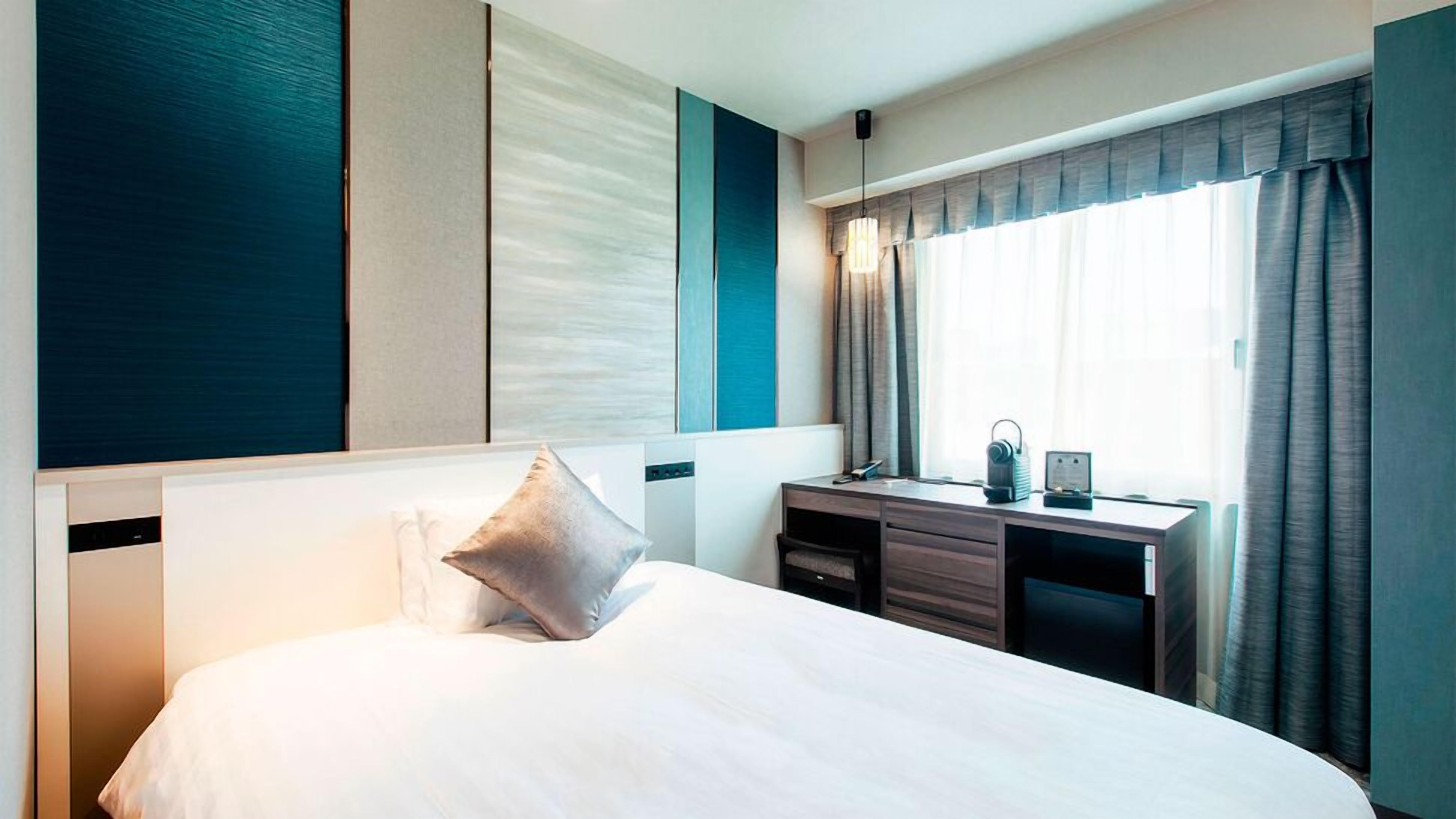 Une chambre d’hôtel comprenant un lit, un bureau, une chaise, une fenêtre avec des rideaux et un mur au décor texturé.