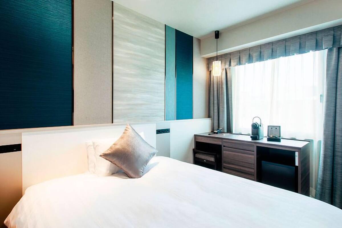 Ett hotellrum med en säng, ett skrivbord, en stol, ett fönster med gardiner och en vägg med ett mönstrat utseende.