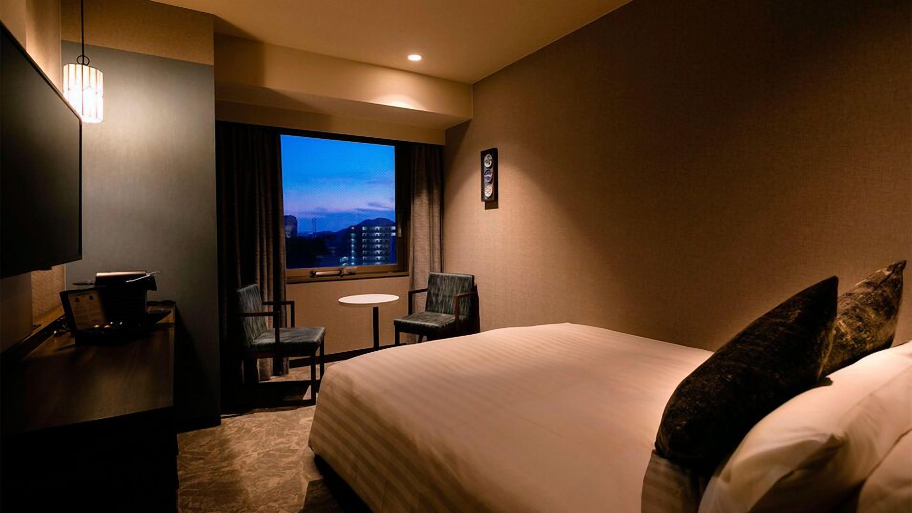 Une chambre d’hôtel avec un lit, un bureau, une chaise, une petite table et une fenêtre donnant sur la ville.