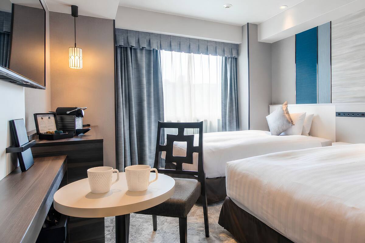 Ett hotellrum med två sängar, ett skrivbord med en stol, ett litet bord med två koppar och ett fönster med gardiner.
