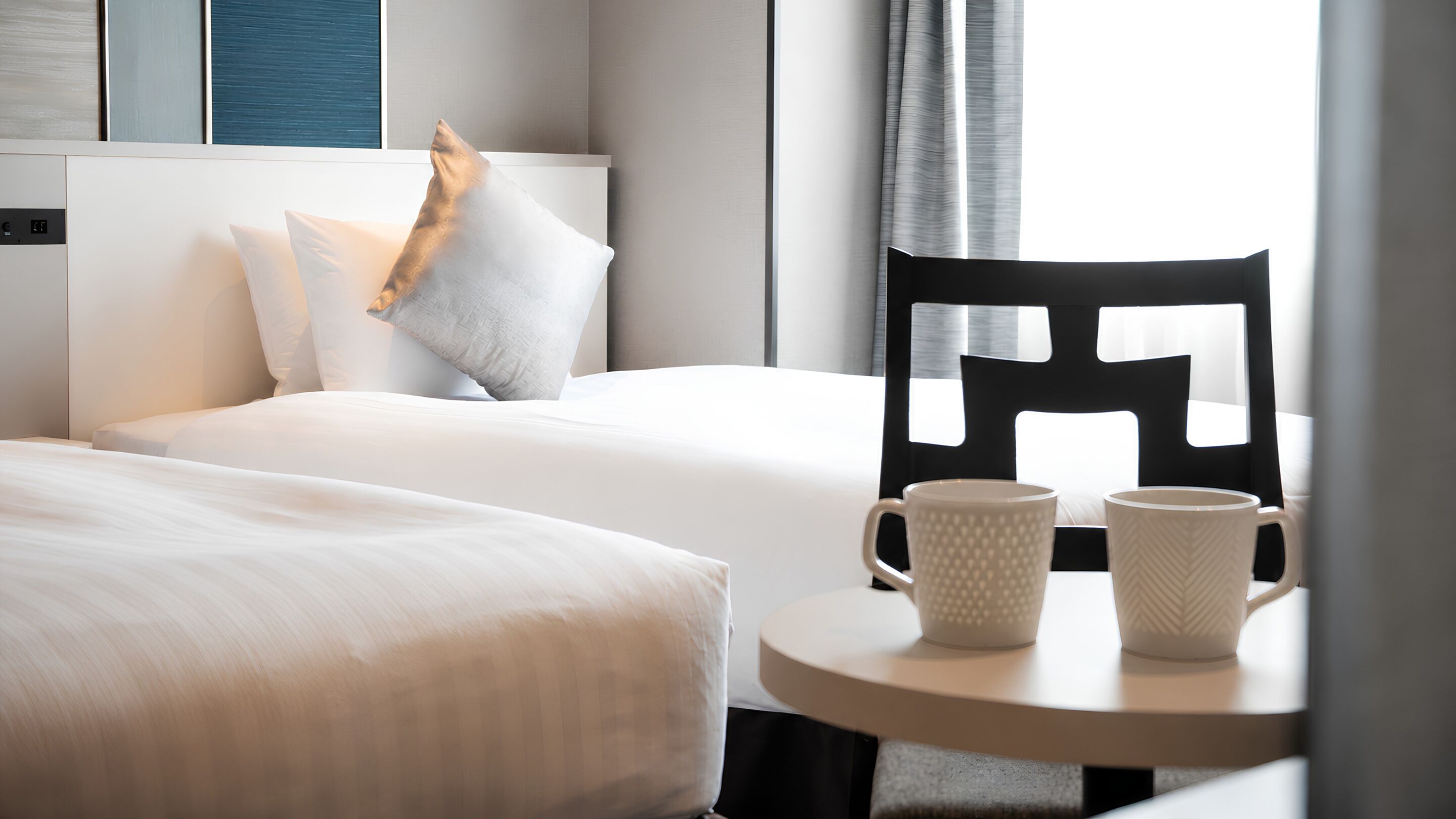 A hotel room with a bed, two pillows, a chair, and two mugs on a small table.