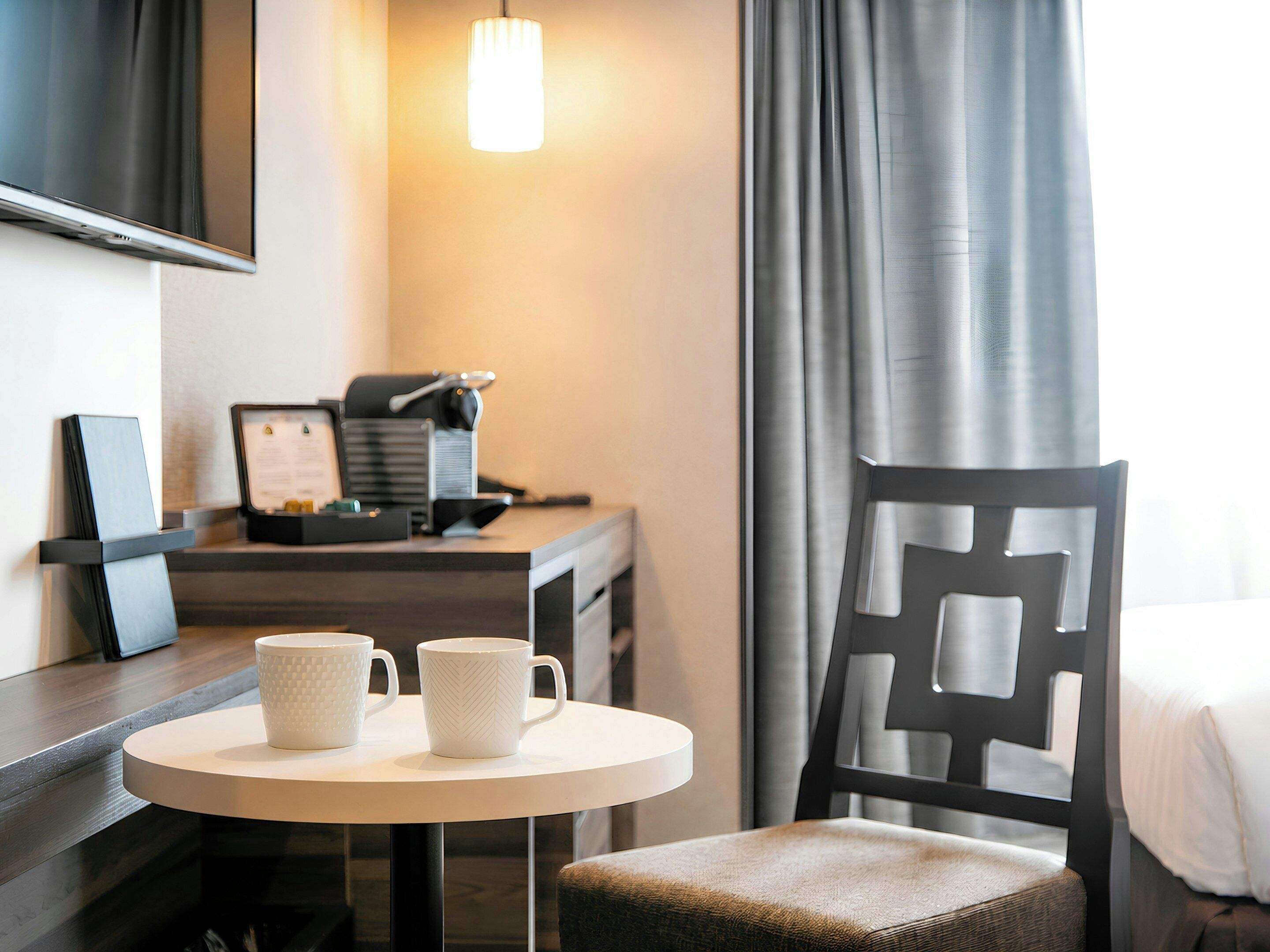 Habitación de hotel con escritorio, dos tazas, lámpara, silla, cama y ventana con cortinas.