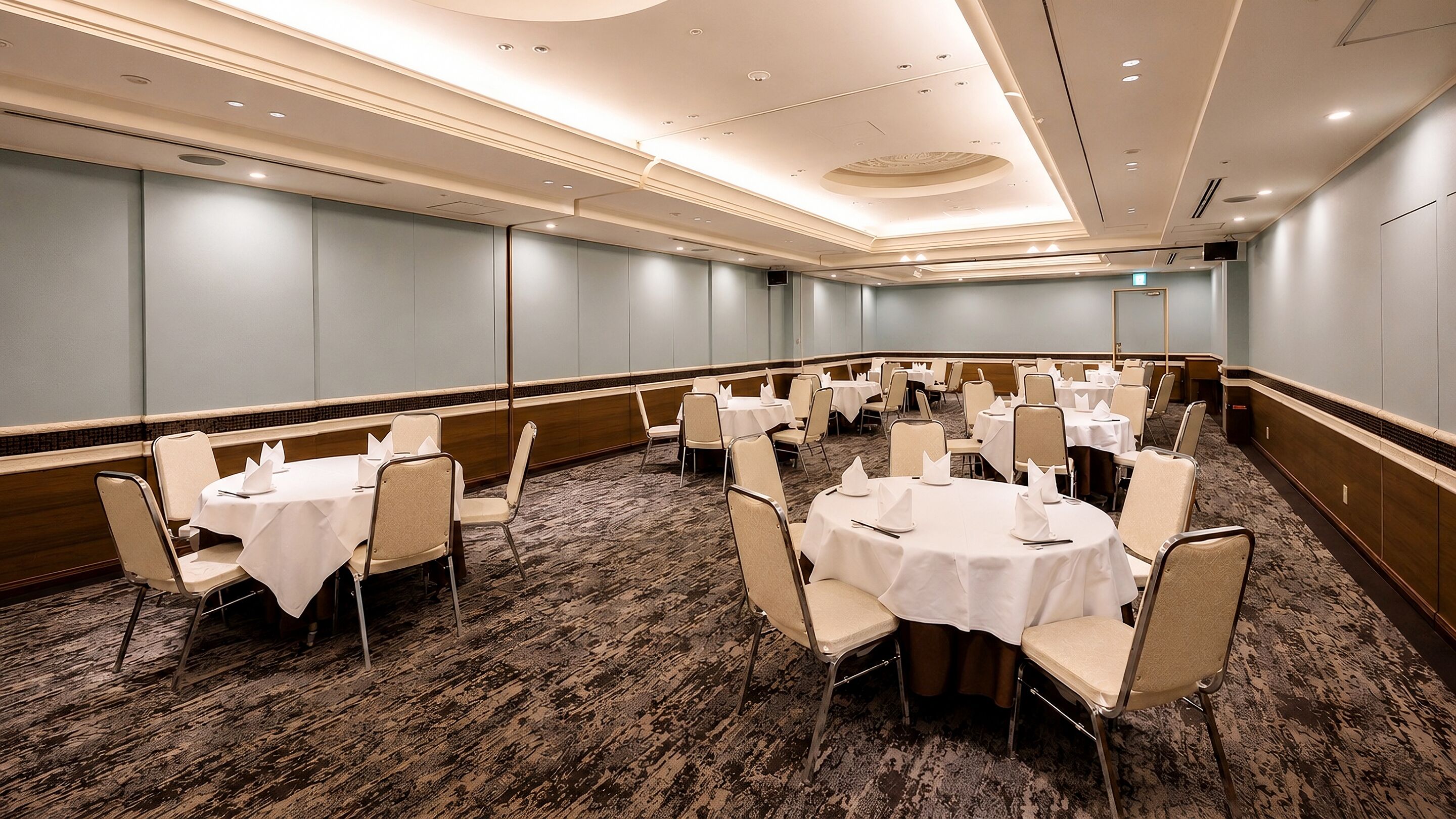 A conference room with multiple round tables and chairs, carpeted flooring, and a high ceiling with recessed lighting.