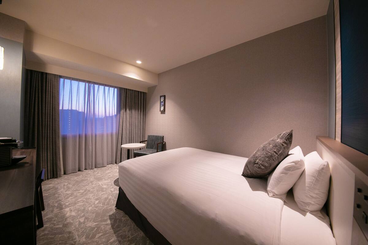 Ett hotellrum med en stor säng, ett litet bord, en stol och utsikt över staden genom fönstret.