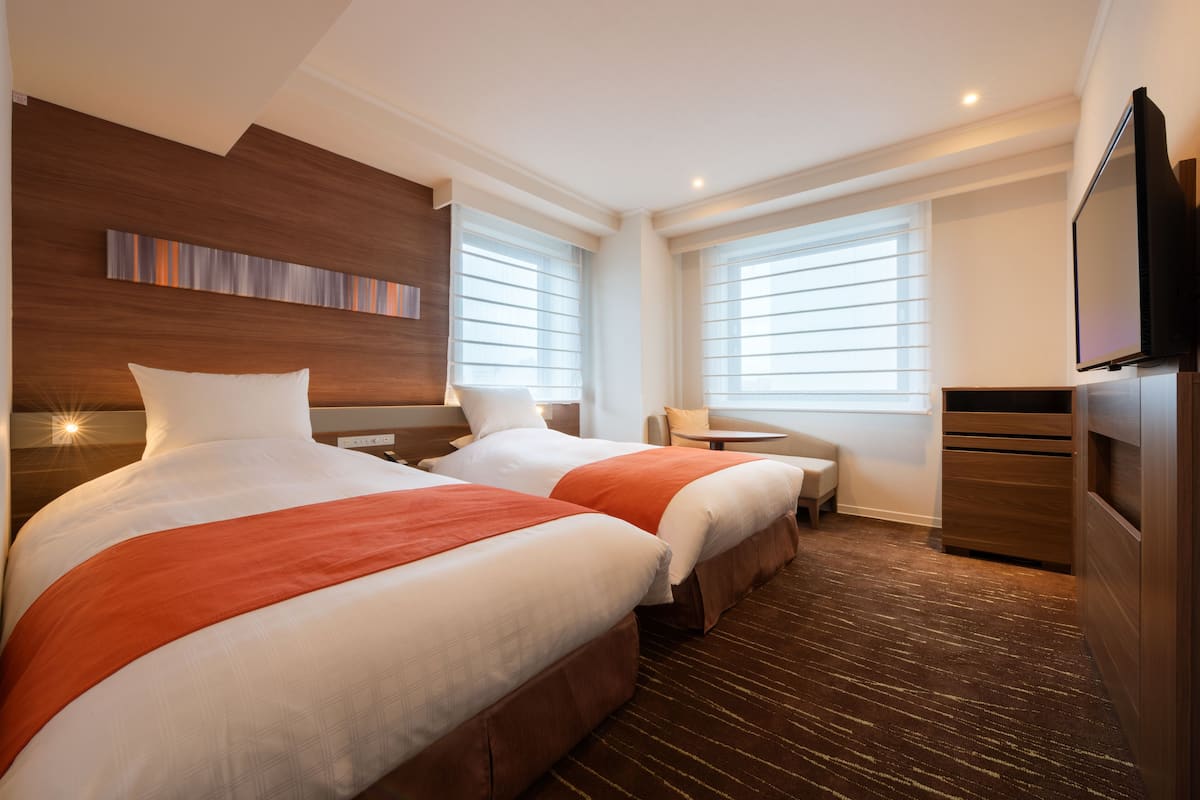 A hotel room with two beds, a TV, a small table, and a window with blinds.