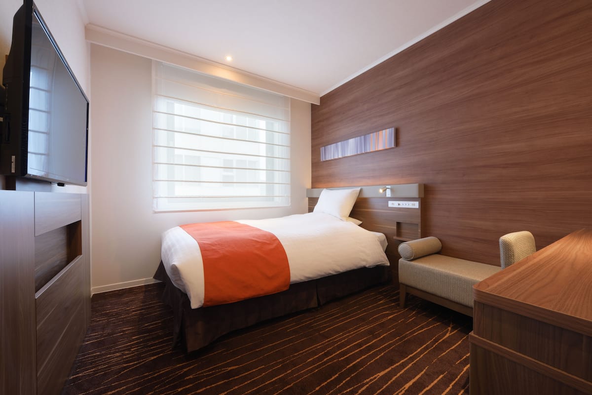 A hotel room with a bed, a TV, a desk, and a window with blinds.