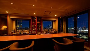 Un bar con una barra larga y numerosas botellas, ventanas amplias con vistas a la ciudad, y dos sillas al frente.