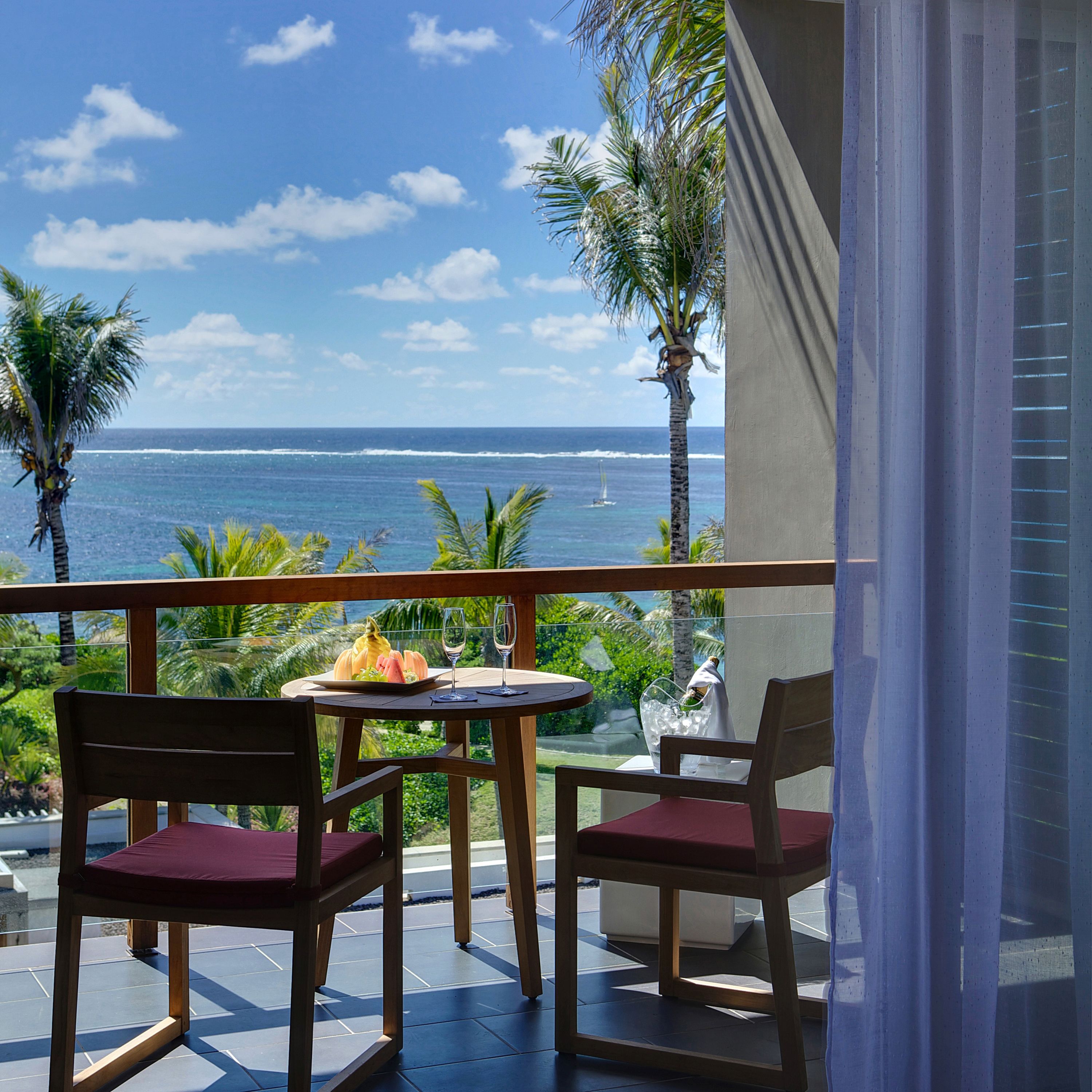 Un balcon avec deux chaises en bois et une table, d’où l’on peut admirer une vue sur l’océan et les palmiers.