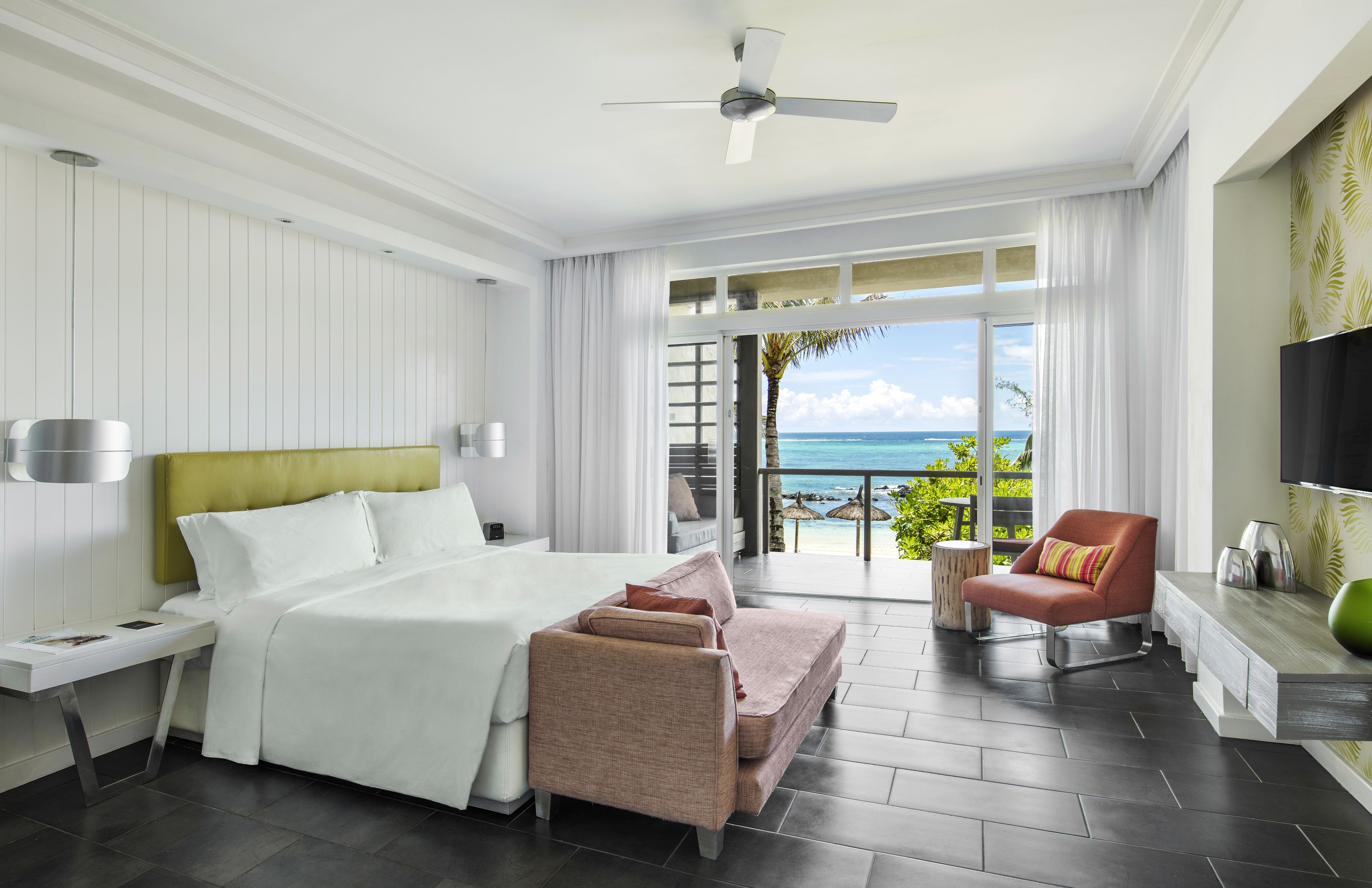 A modern hotel room with a large bed, a seating area with a chair, a TV, and a balcony with a view of the ocean.