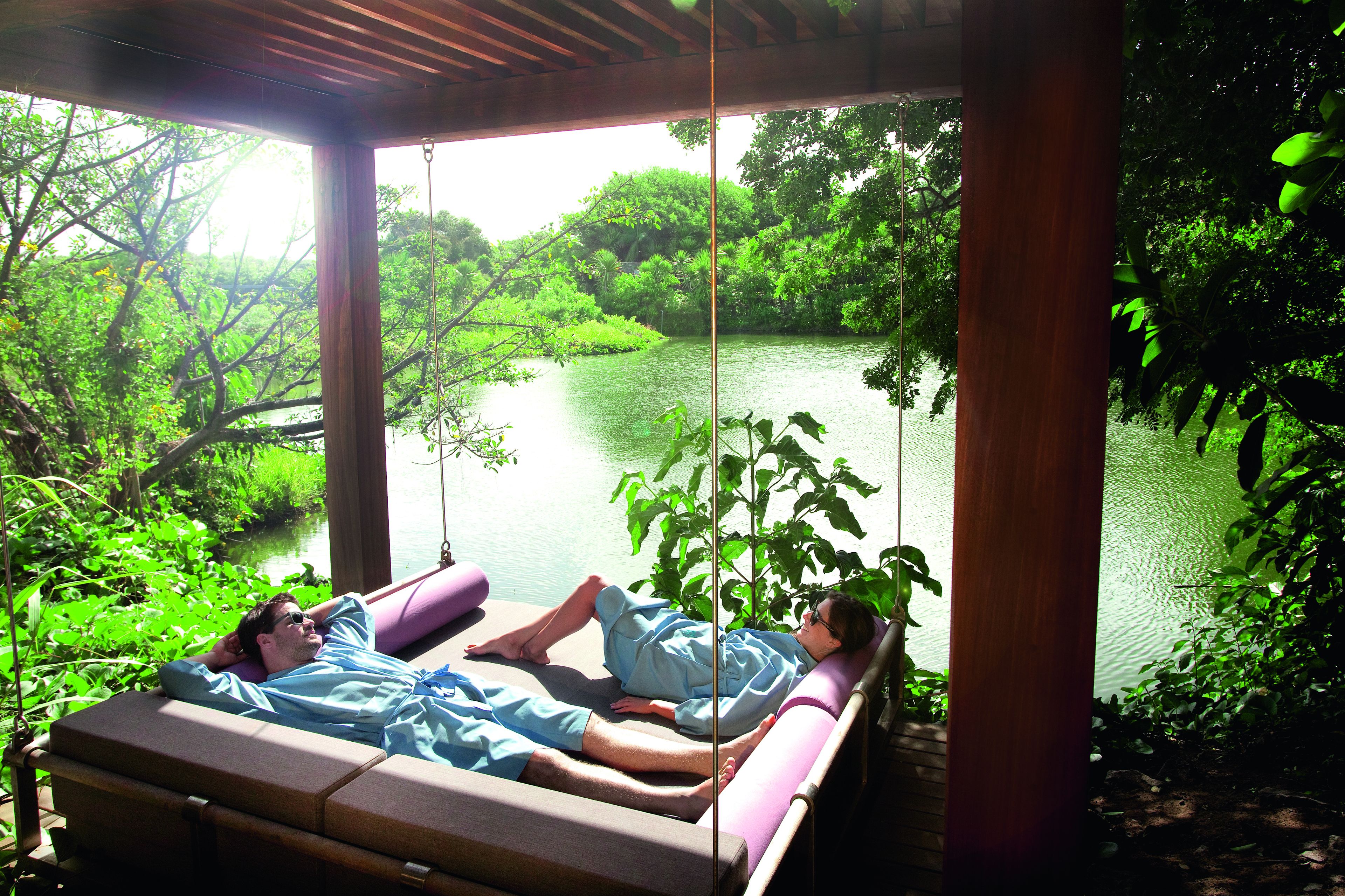 A man lying on a cushioned bench with a view of a lake and greenery.