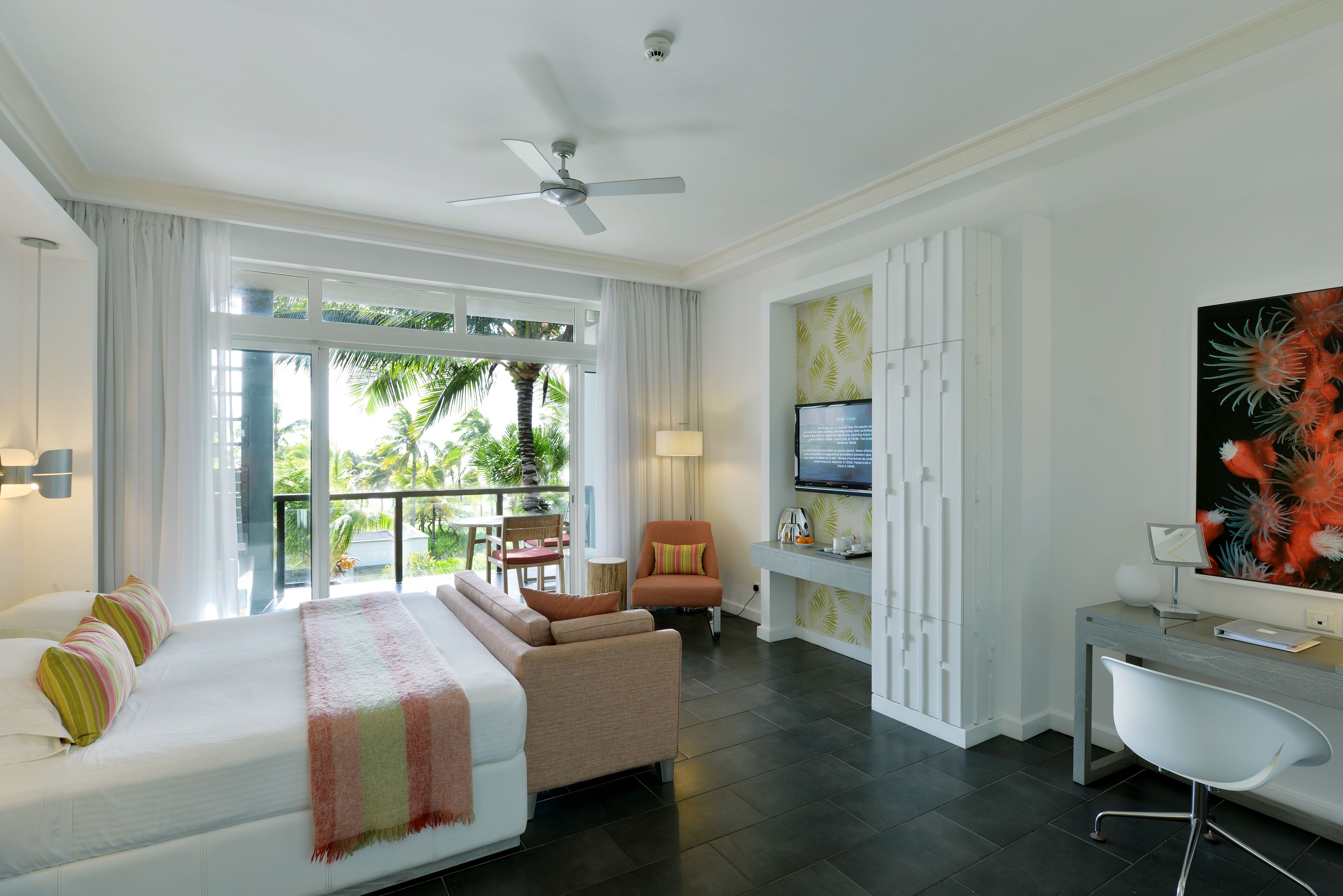 A modern hotel room with a large bed, a desk, a chair, and a balcony with a view of palm trees.