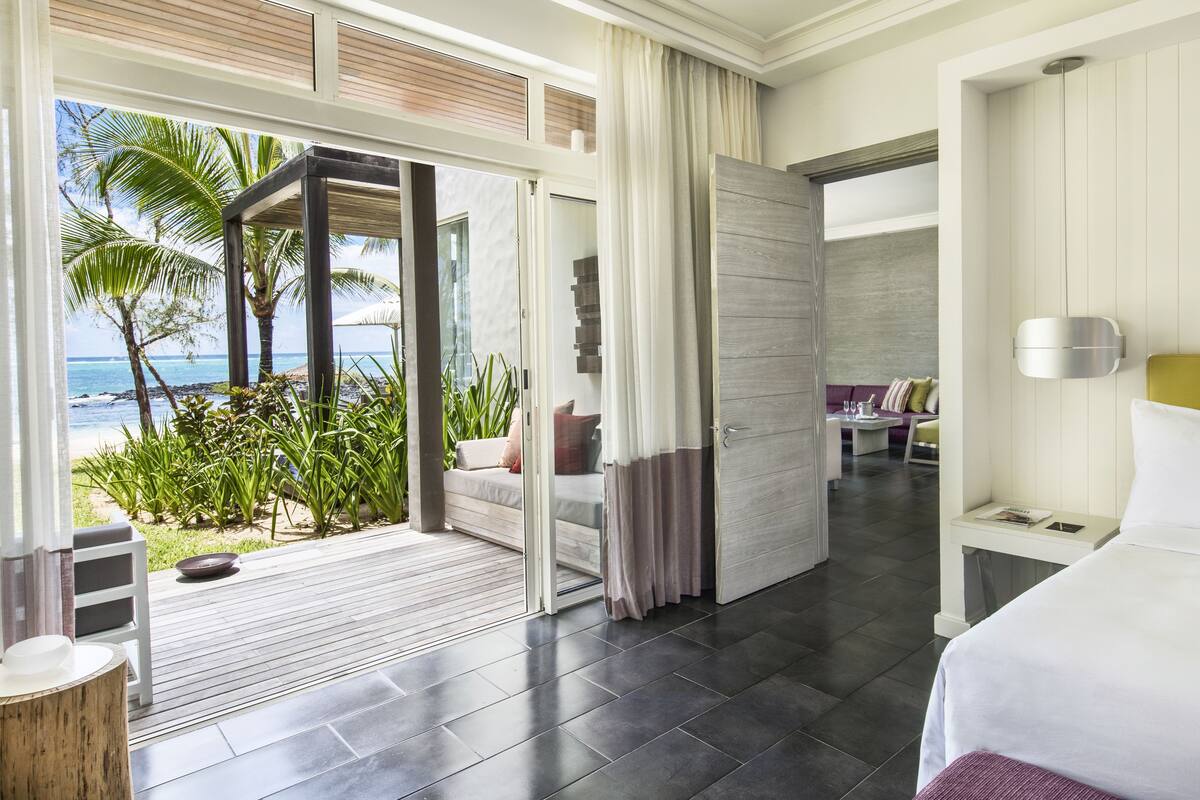 Une chambre d’hôtel dotée d’une grande porte-fenêtre coulissante donnant sur un balcon avec vue sur l’océan et les palmiers. La chambre comprend un lit, une table de chevet et un coin salon avec un canapé et une petite table.
