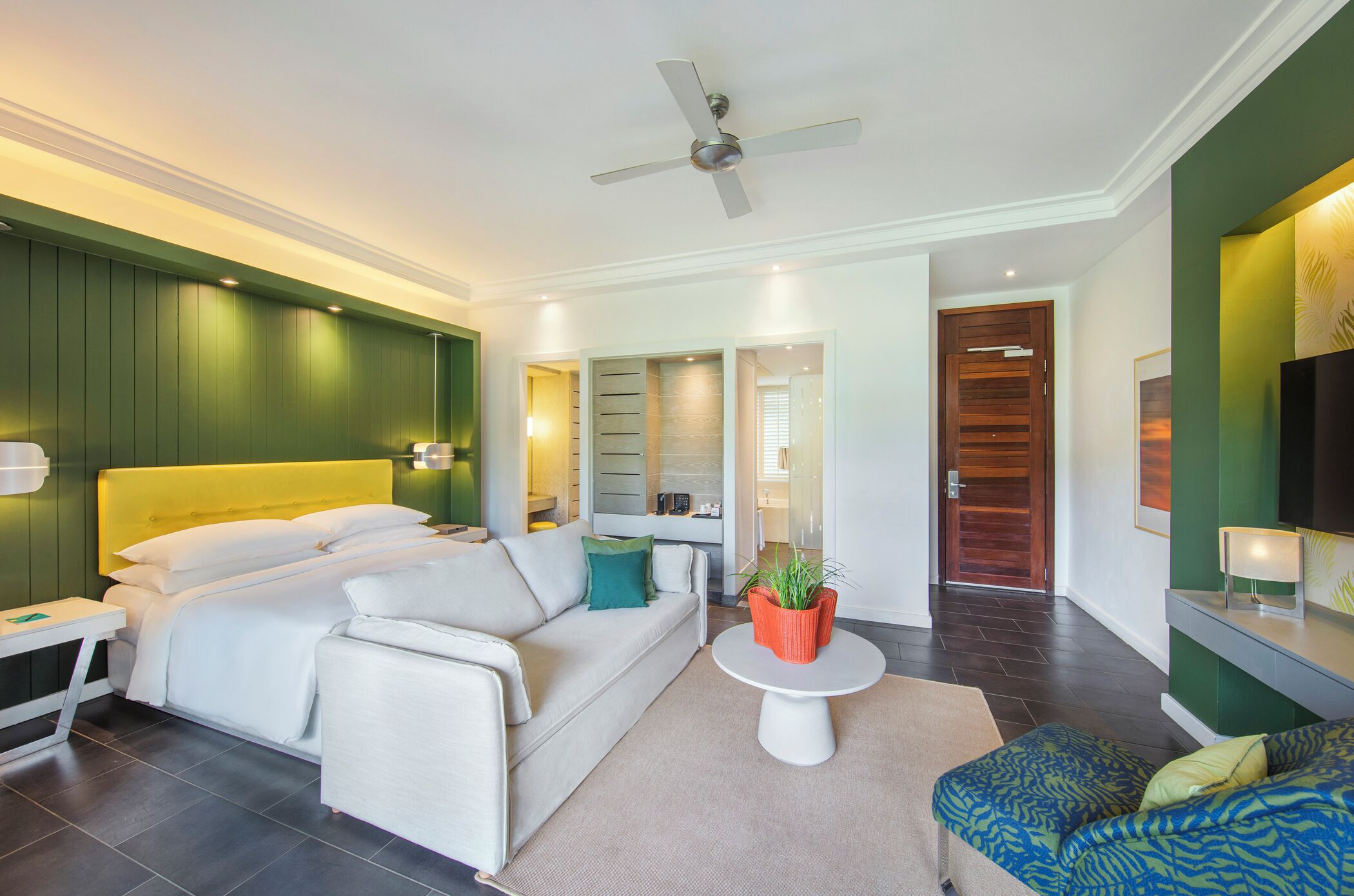 A modern hotel room with a bed, a sofa, a small table with a plant, and a ceiling fan.