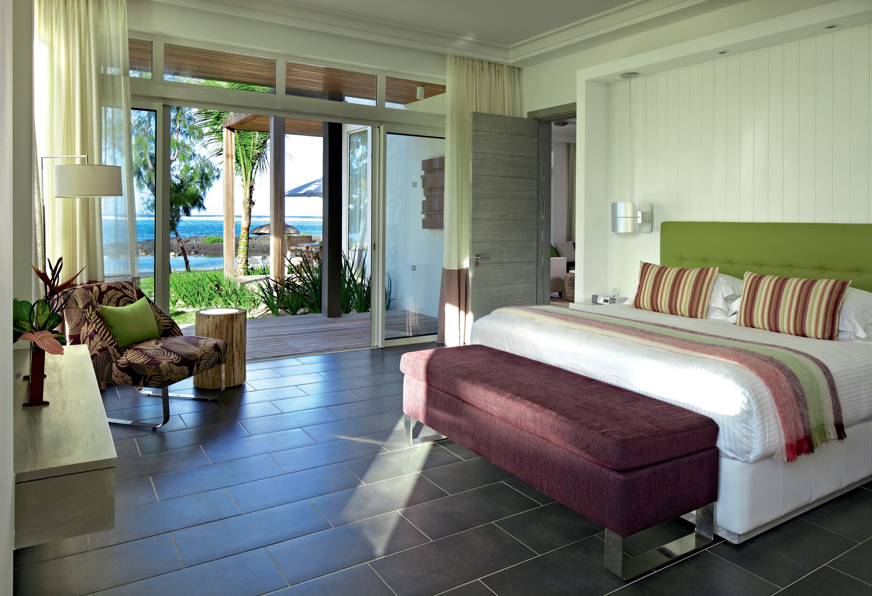 A modern bedroom with a large bed, a bench, a sofa, and a view of the ocean.